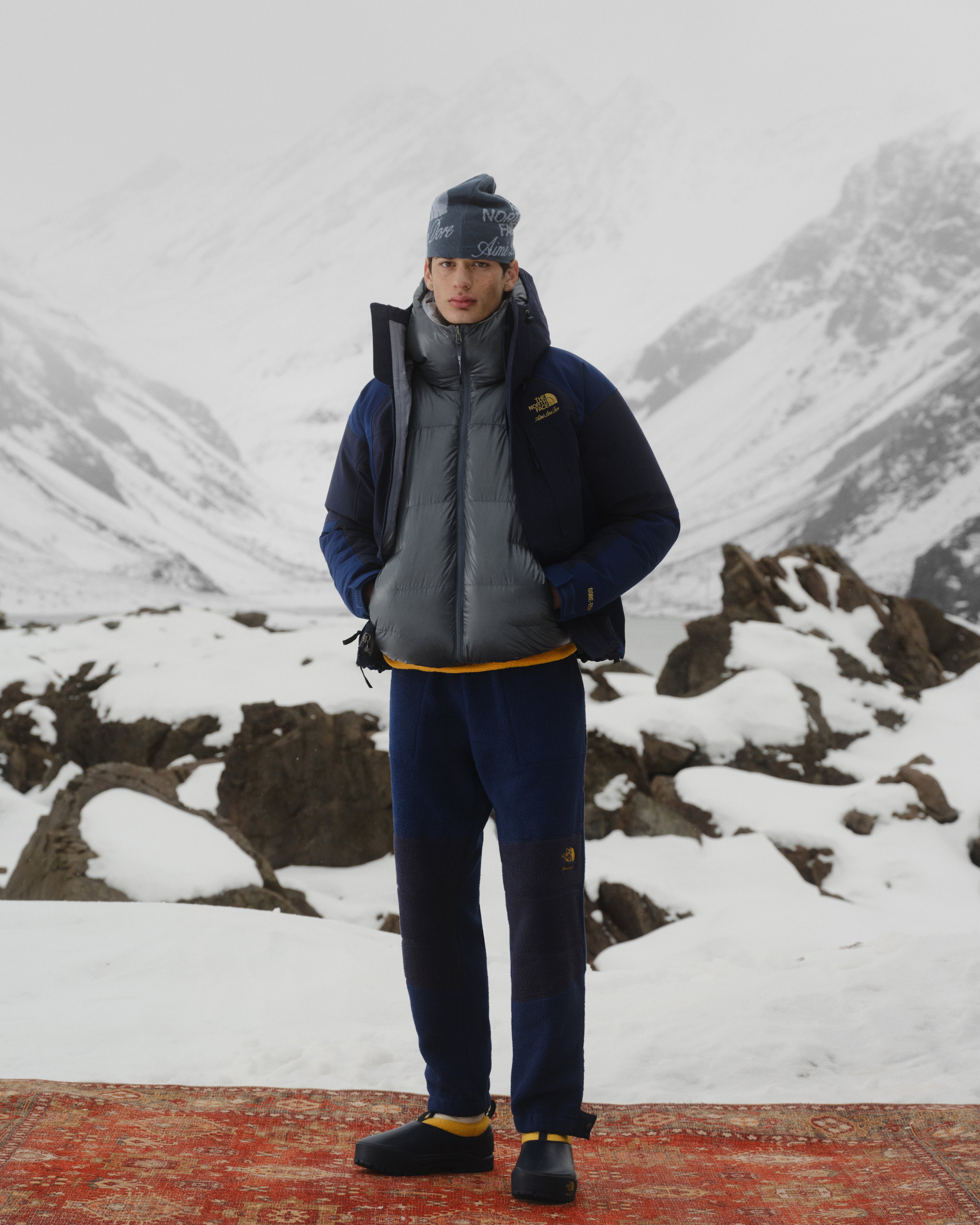 Person in winter clothes wears ALD / The North Face Base Camp Mule on a red rug amid snowy mountains and rocky peaks.