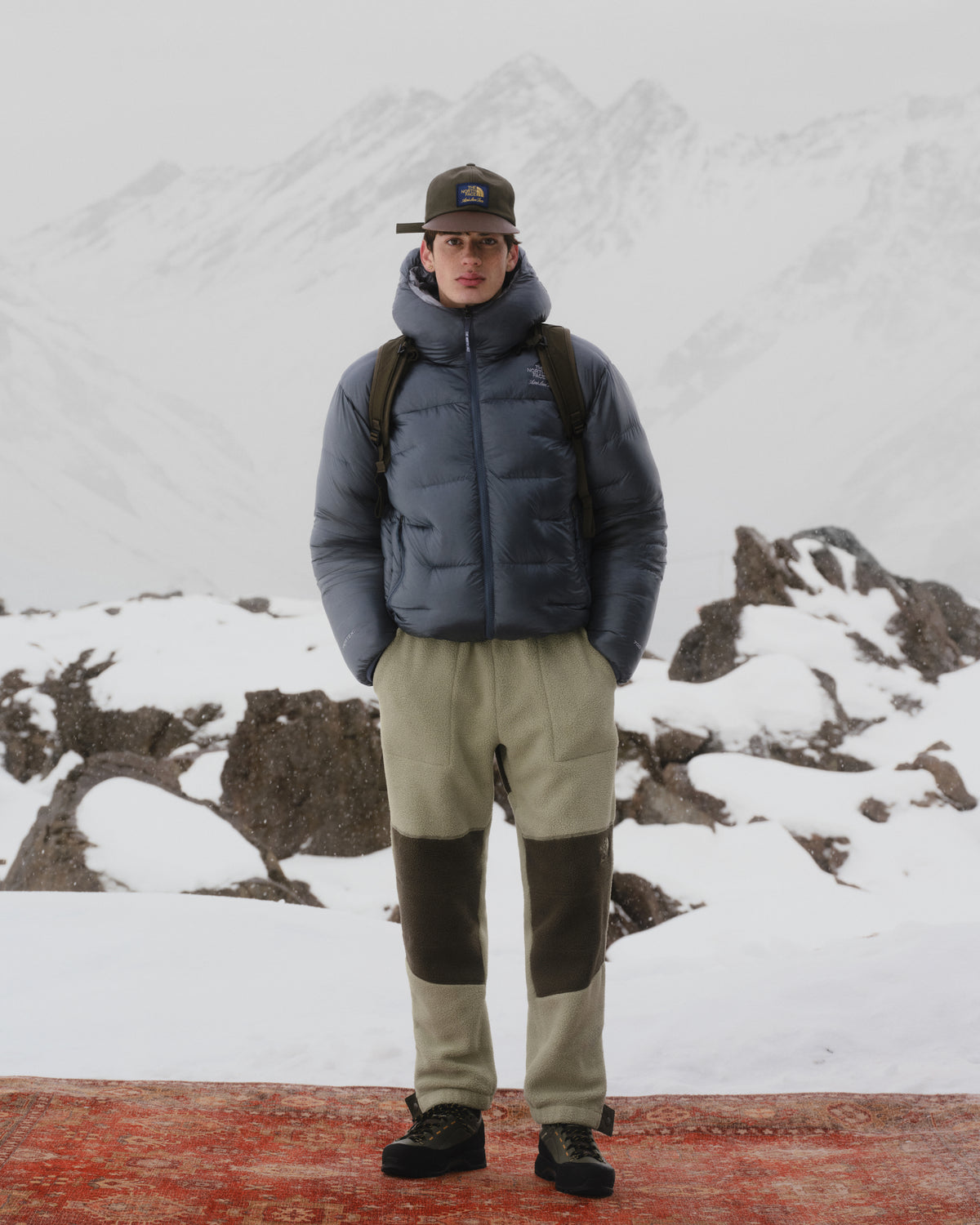 A person wears the ALD / The North Face Reversible Pertex® Nuptse Jacket on a red rug, snowy mountains behind.
