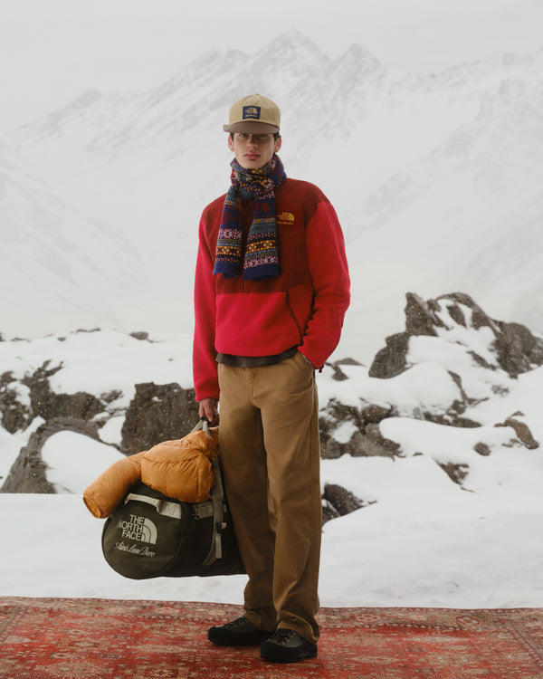 A man with the ALD / The North Face Fair Isle Scarf stands on snow, holding a duffel bag, mountains in the background.