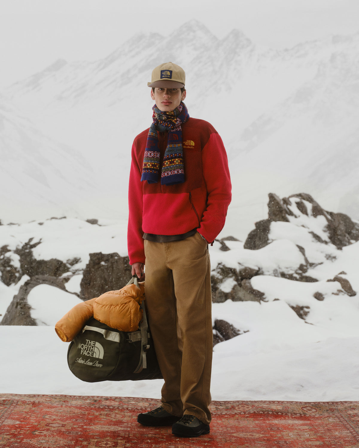 A man in winter gear stands on a rug with the ALD/The North Face Base Camp Duffel, snowy peaks behind him.