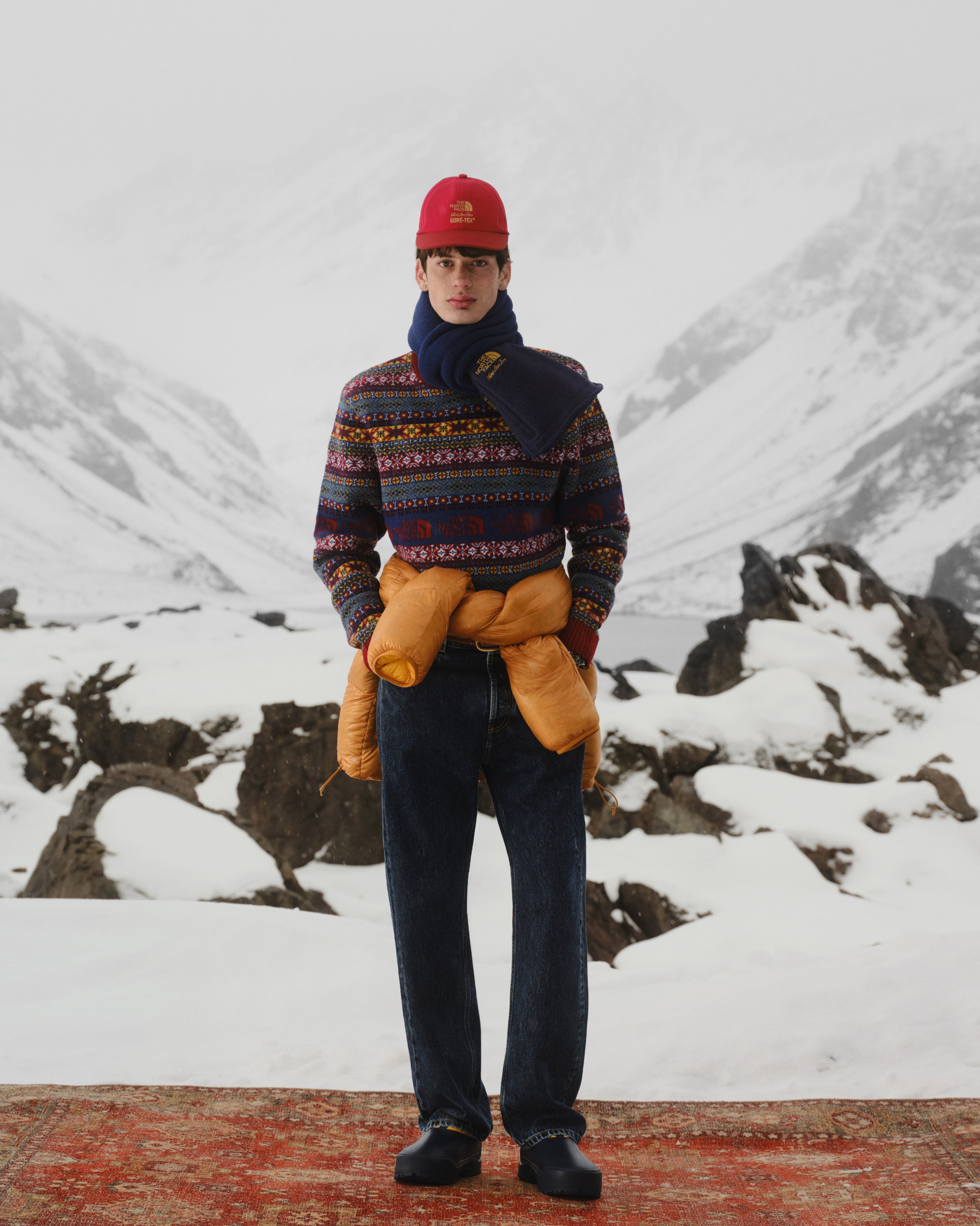A man in winter clothes stands on a rug in snow, wearing the ALD/The North Face Denali Fleece Scarf.