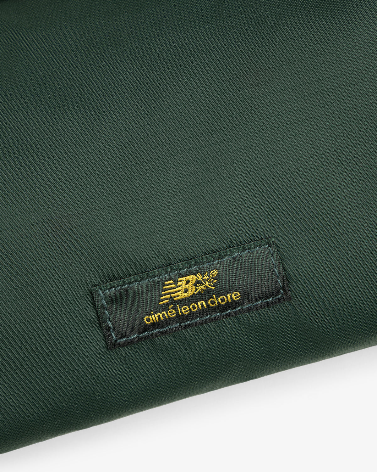 Close-up of a green ALD / New Balance Nylon Sling Pouch with a black patch featuring gold New Balance aimé leon dore text.
