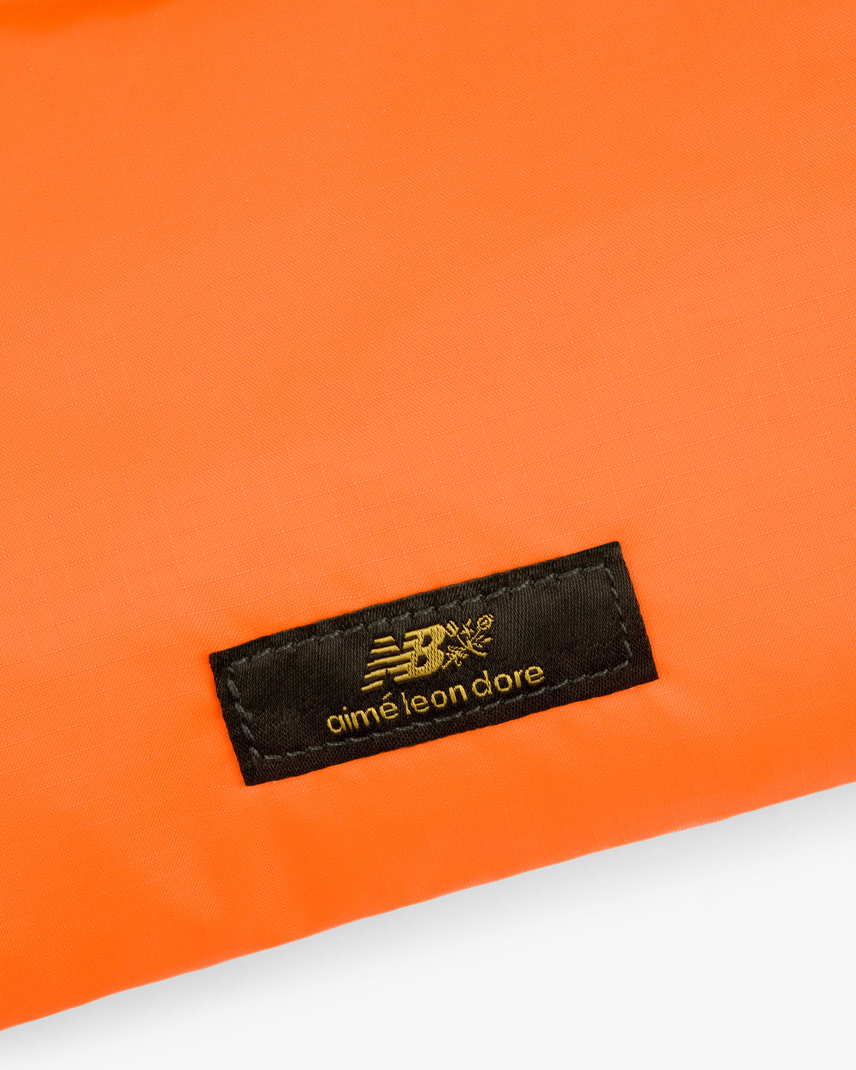 Black ALD / New Balance label sewn onto bright orange Nylon Sling Pouch fabric.