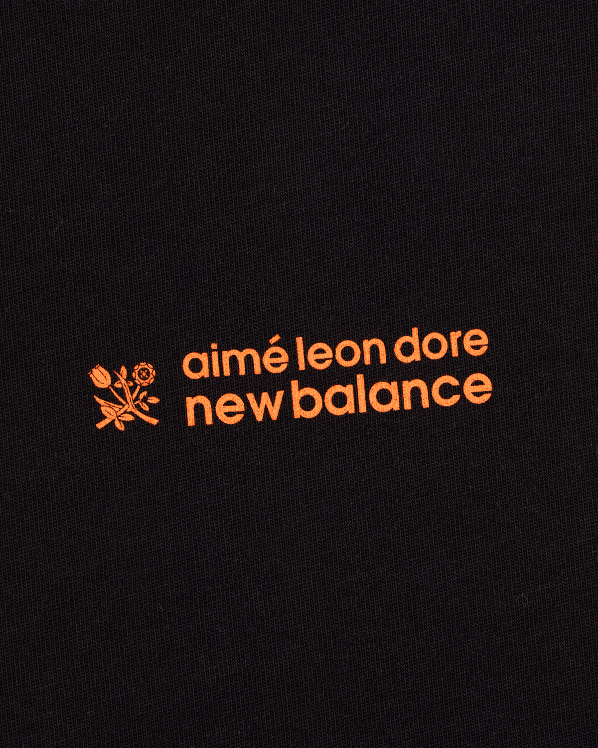 ALD / New Balance Running Bib Tee features an orange logo with a flower graphic on black fabric.