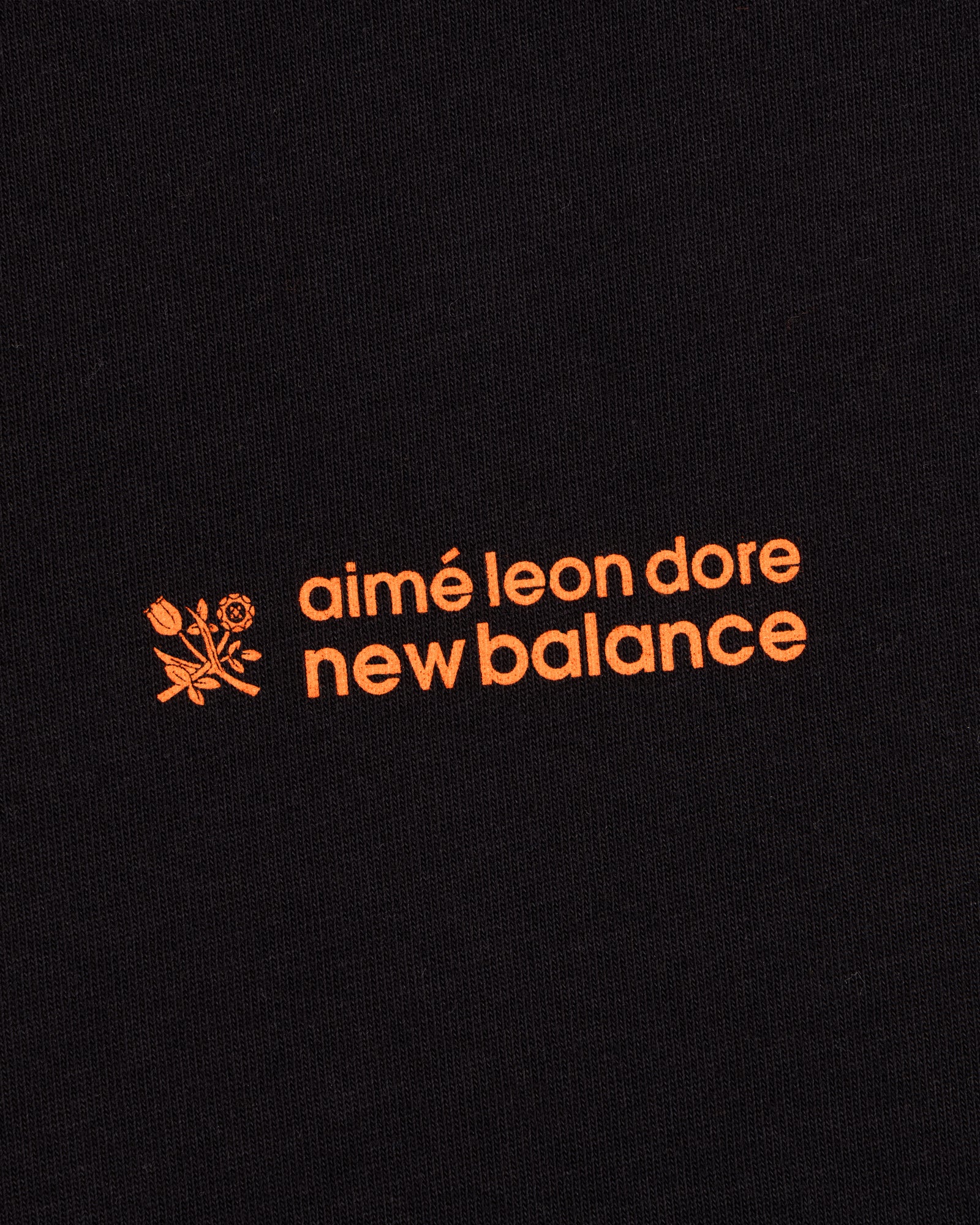 ALD / New Balance Running Bib Tee features an orange logo with a flower graphic on black fabric.