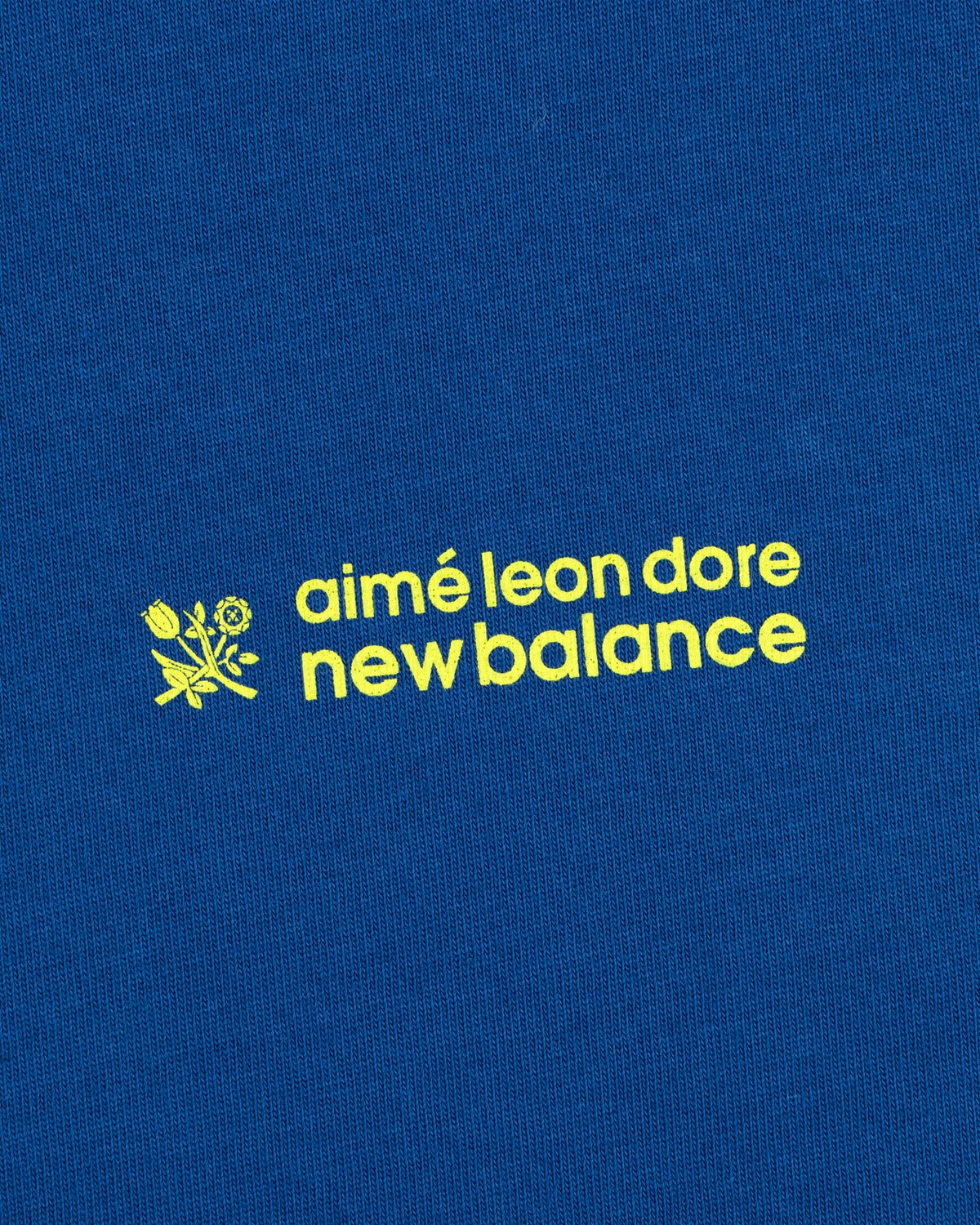 Yellow "ALD / New Balance Running Bib Tee" text and small flower graphic on a blue fabric background.