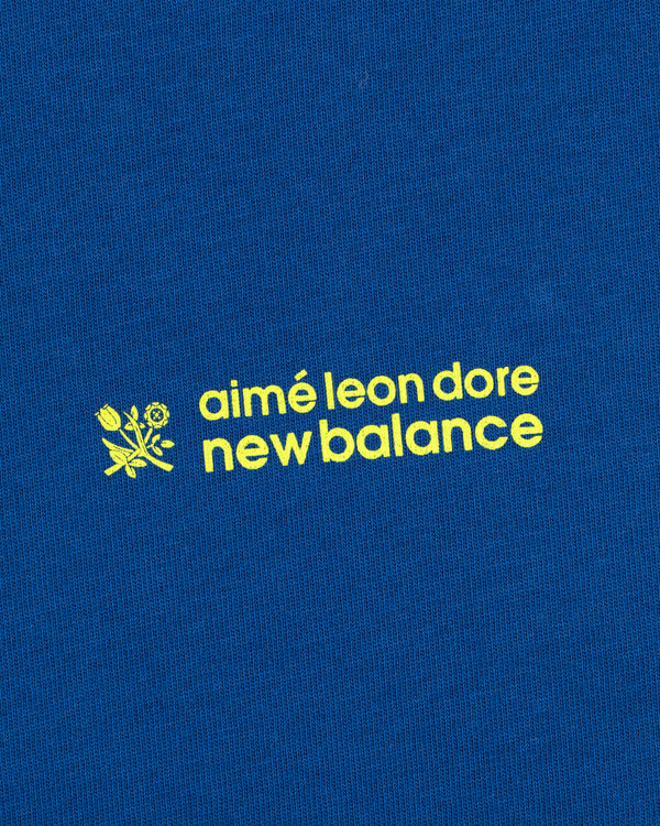 Yellow "ALD / New Balance Running Bib Tee" text and small flower graphic on a blue fabric background.