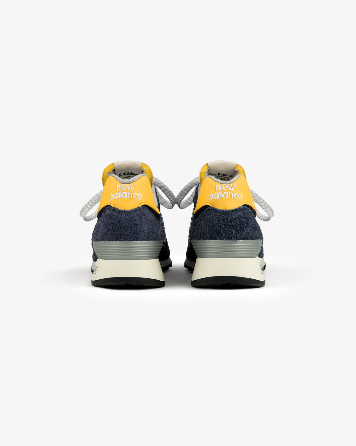 The ALD / New Balance Made in USA 1300 sneakers in navy and yellow, viewed from the back on a white background.