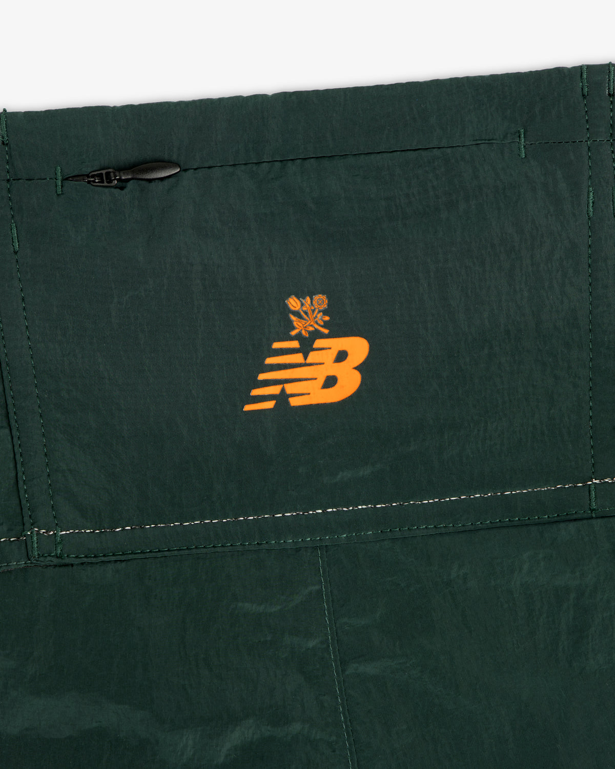 Close-up of dark green ALD / New Balance Lightweight Nylon Warmup Pant with orange logo and small flower above it.