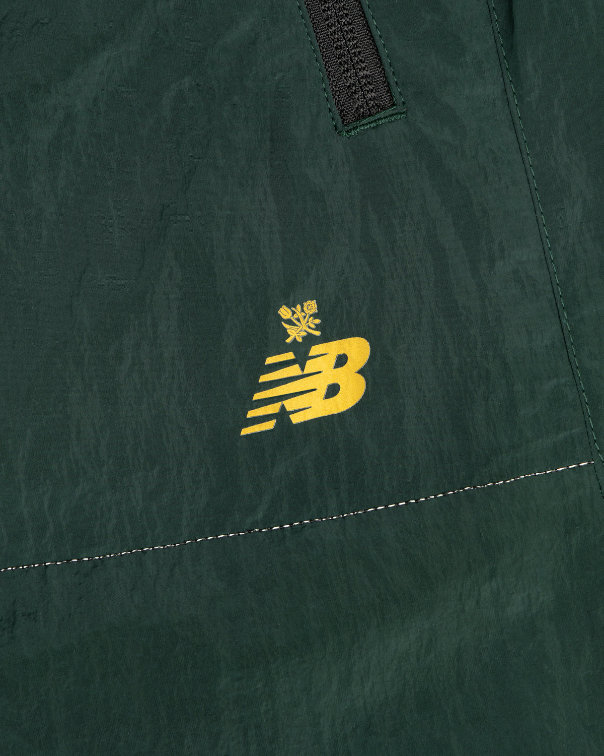 Close-up of ALD / New Balance Lightweight Nylon Warmup Pant in green with yellow NB logo, crown, seam, and zipper.