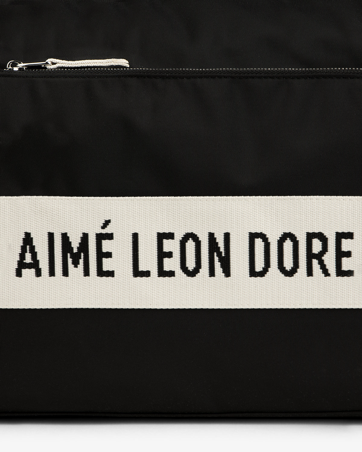 Black nylon duffle bag with a cream stripe featuring bold black AIMÉ LEON DORE text.