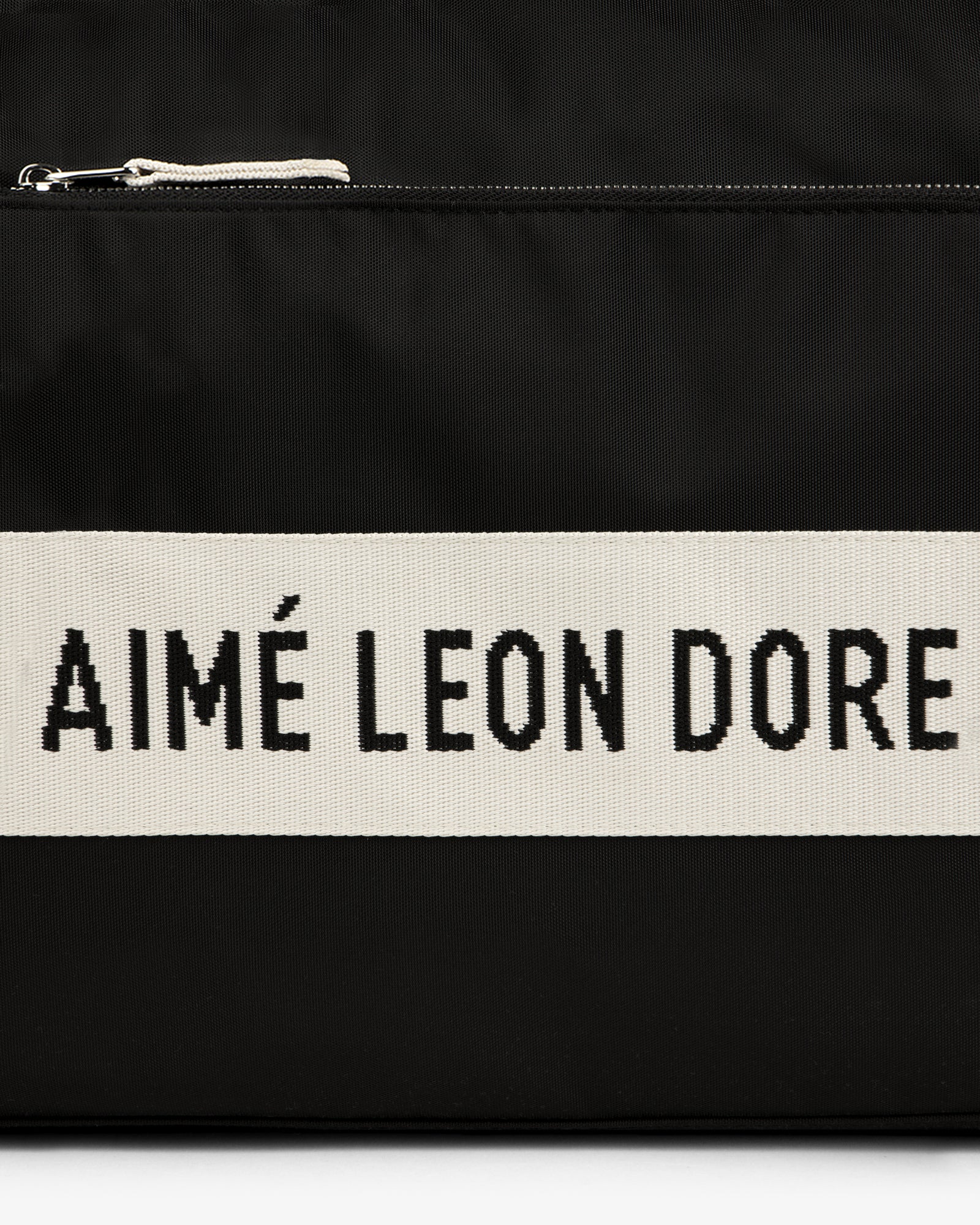 Black nylon duffle bag with a cream stripe featuring bold black AIMÉ LEON DORE text.