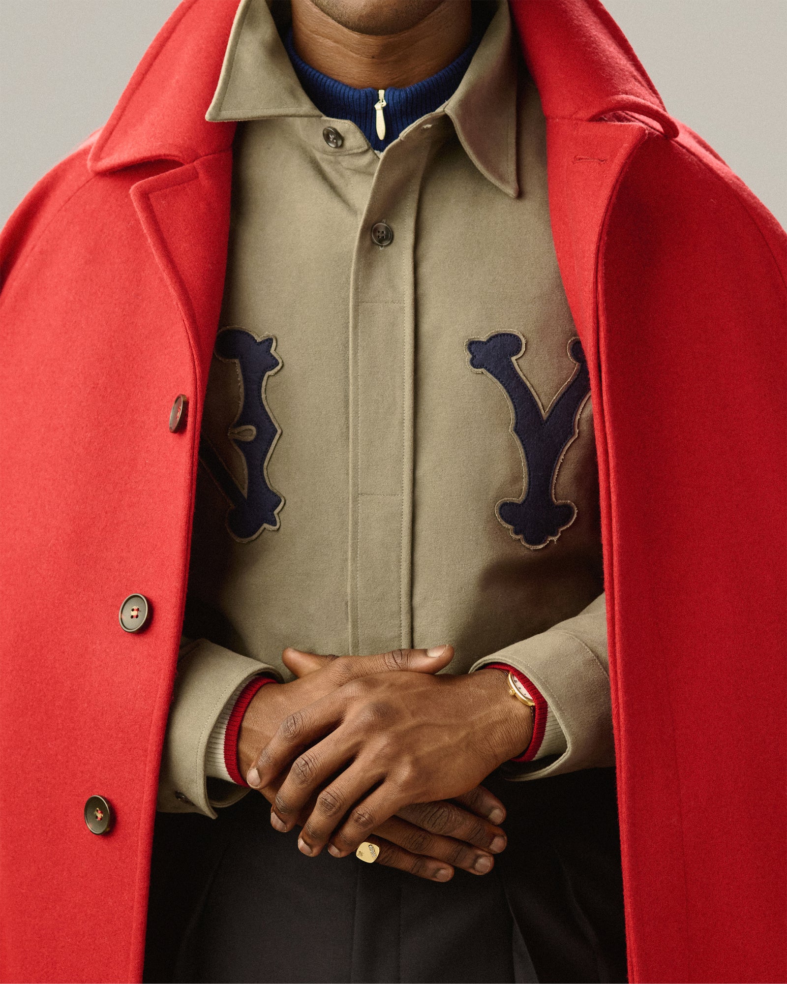 A person in a red coat over a tan shirt, hands clasped, wearing a 14Kt Gold Diamond Signet Ring and a watch.