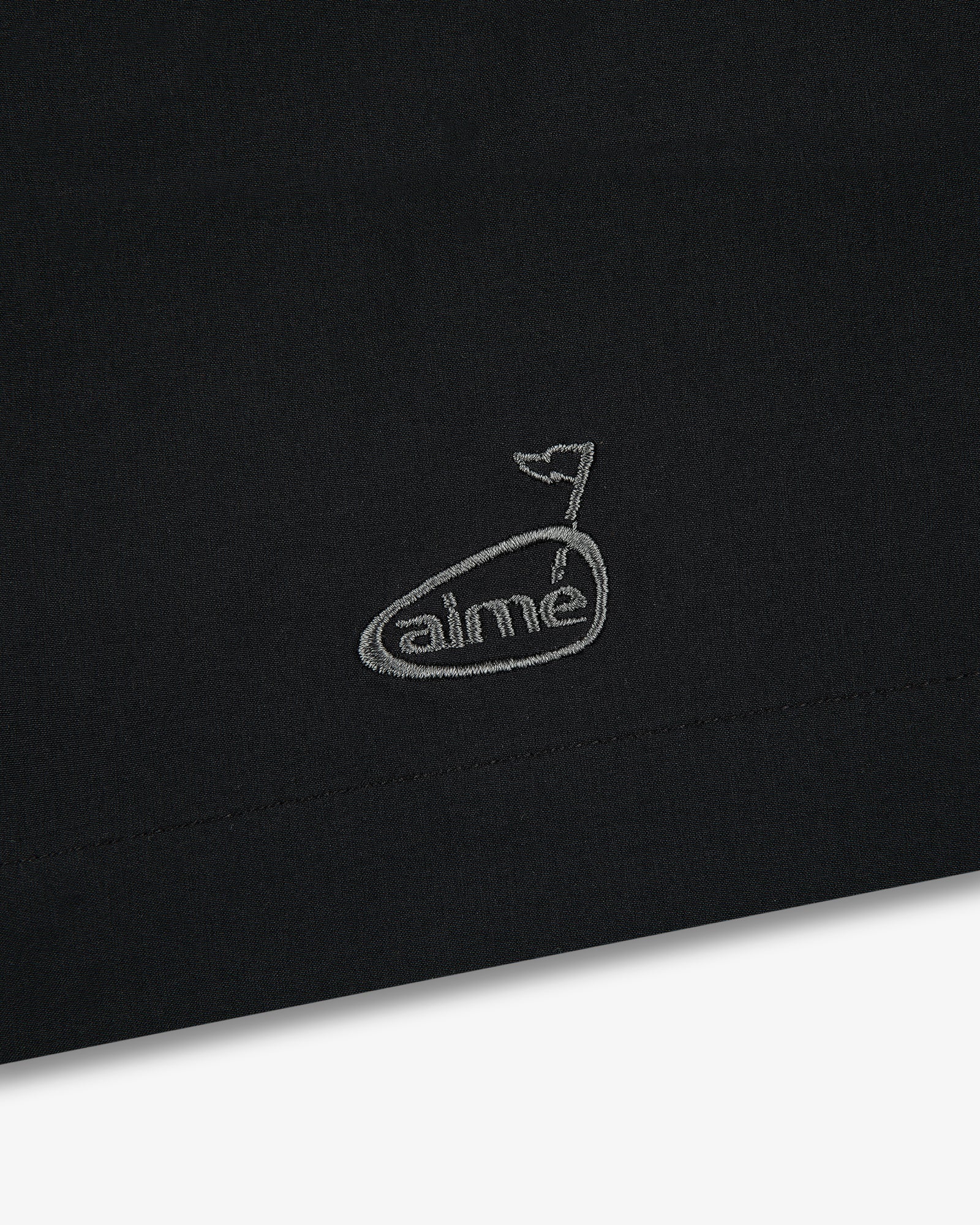 Black fabric with a small, white embroidered whale above “aimé” on the ALD Golf Performance Short.