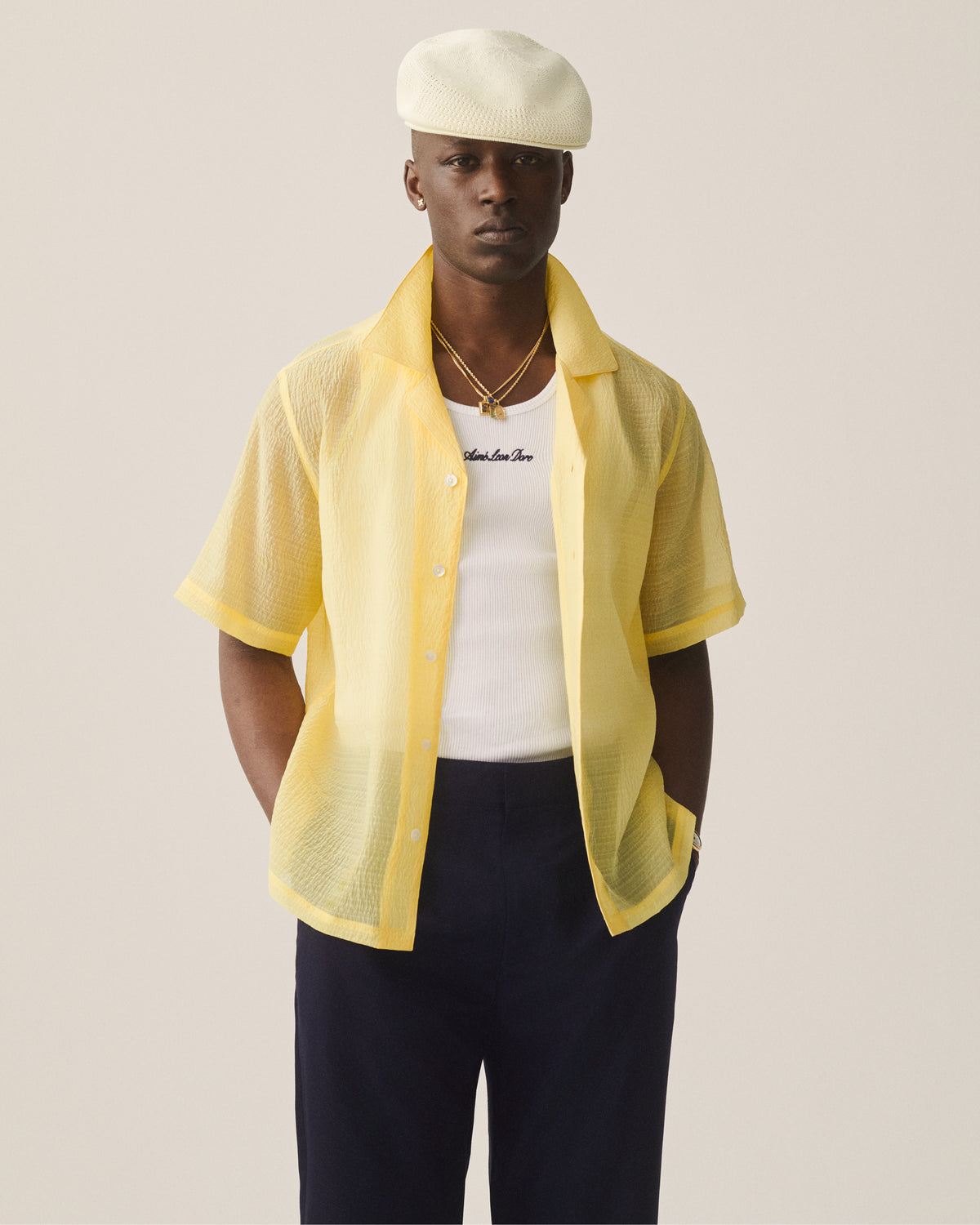A man stands with hands in pockets, wearing a yellow short-sleeve shirt, navy pants, white beret, and Embroidered Logo Tank Top.