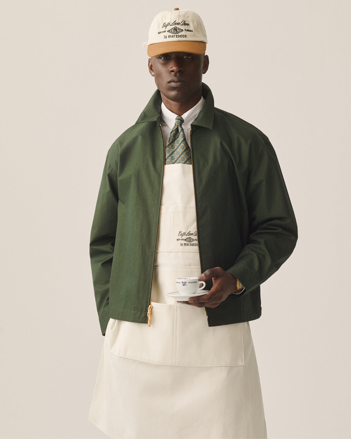 A man in a green jacket and tie wears the Café Leon Dore / La Marzocco Apron, holding a coffee cup and saucer.