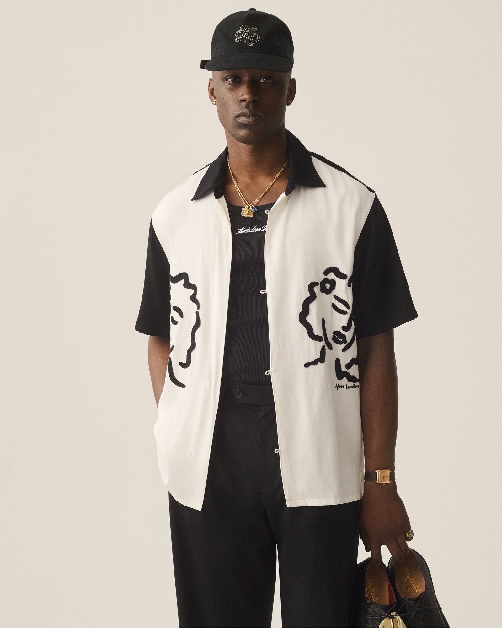 A man in black pants and a black cap wears an Embroidered Logo Tank Top and holds shoes.