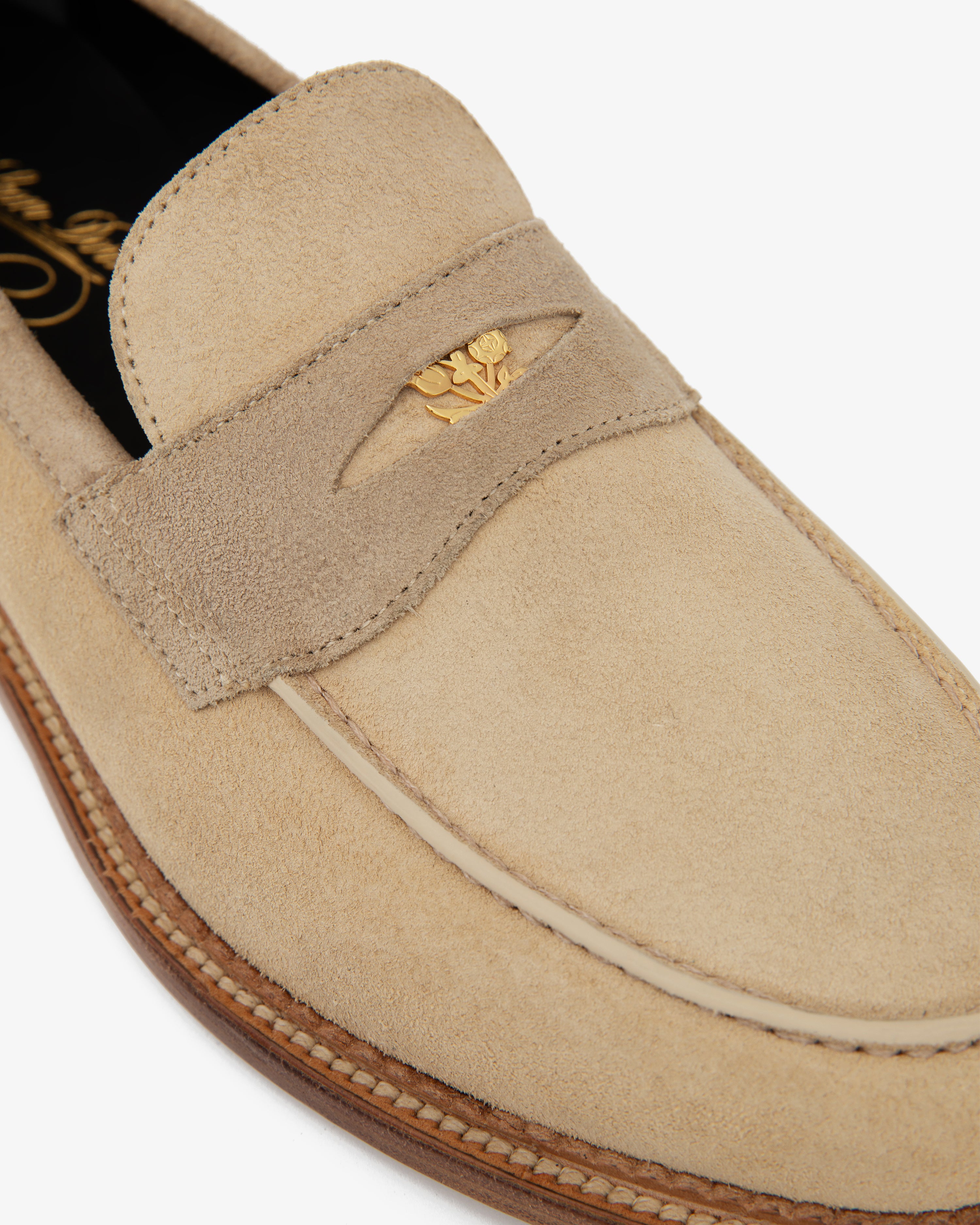 Close-up of the Suede Queens Crest Loafer in beige with gold emblem detail and sole stitching.