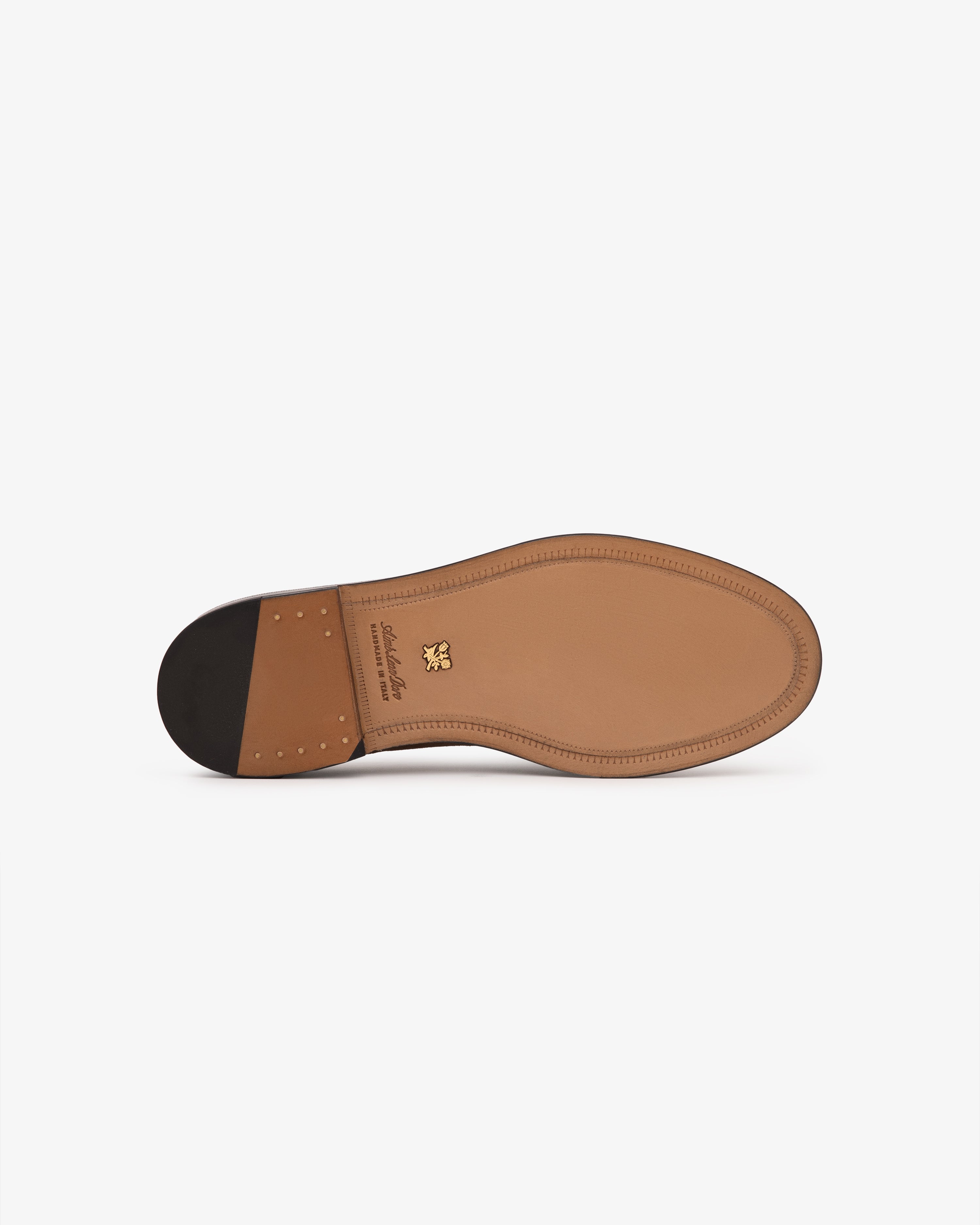 The Suede Queens Crest Loafer features a stitched sole, black heel, and gold logo, shown on a white background.