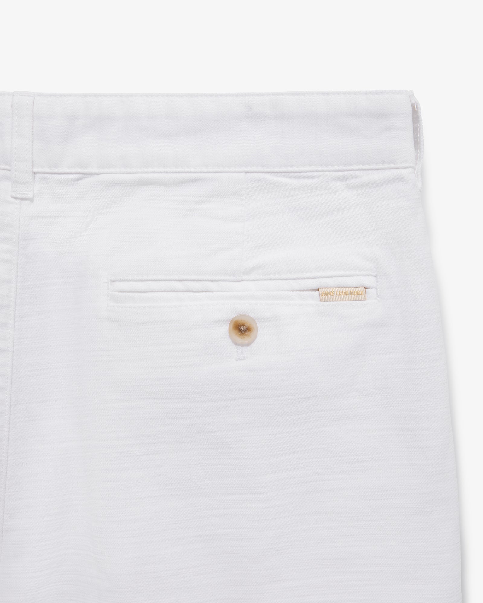 Close-up of Double Pleated Pant’s back pocket with a button and beige brand label above the pocket.