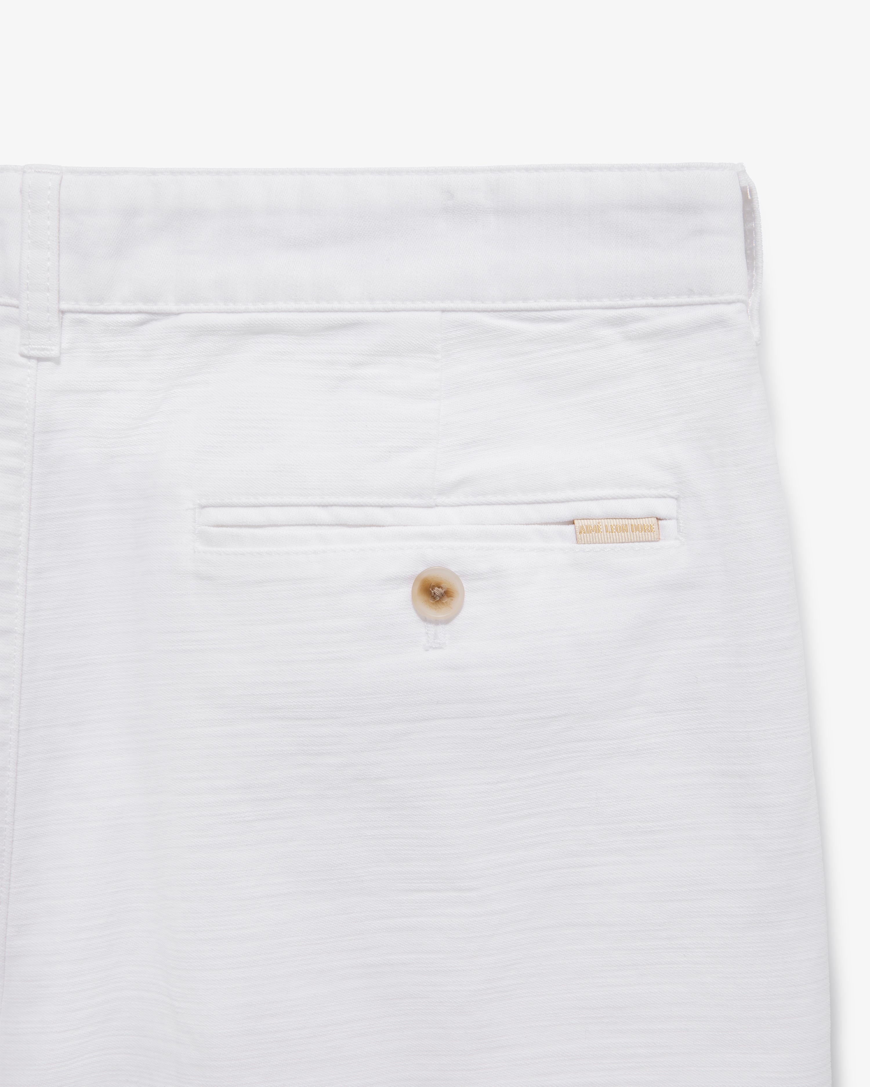 Close-up of Double Pleated Pant’s back pocket with a button and beige brand label above the pocket.