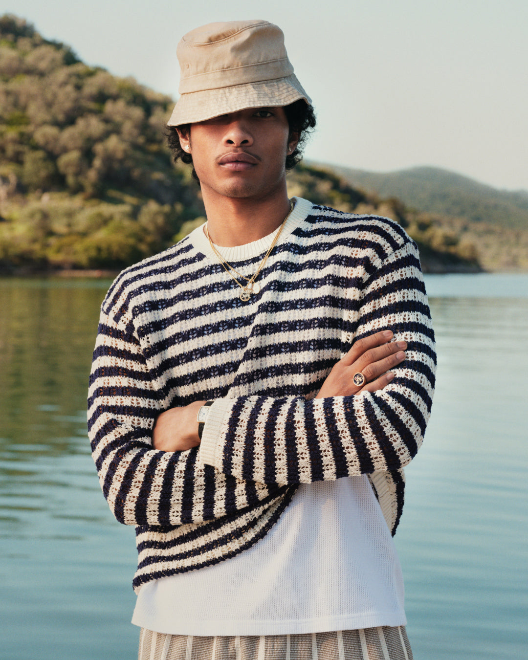 Man wearing a Skáfos Sweater, beige bucket hat, and jewelry stands by water with trees and hills in the background.