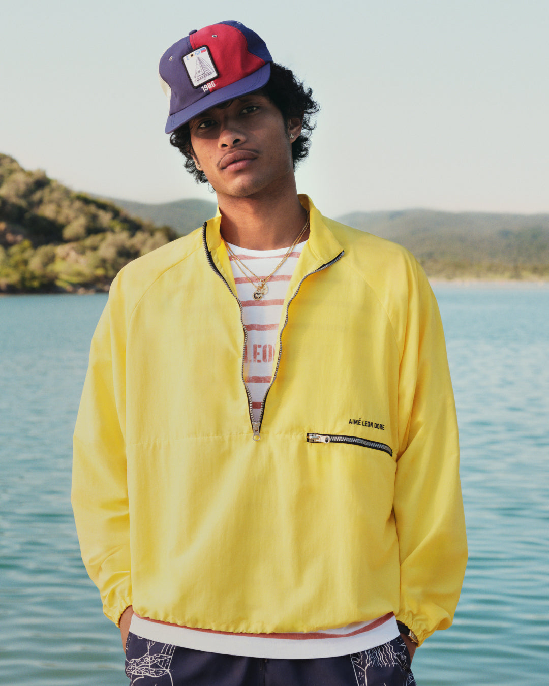 A person in a yellow jacket and Nylon Deck Hat stands by a lake with hills and trees in the background.