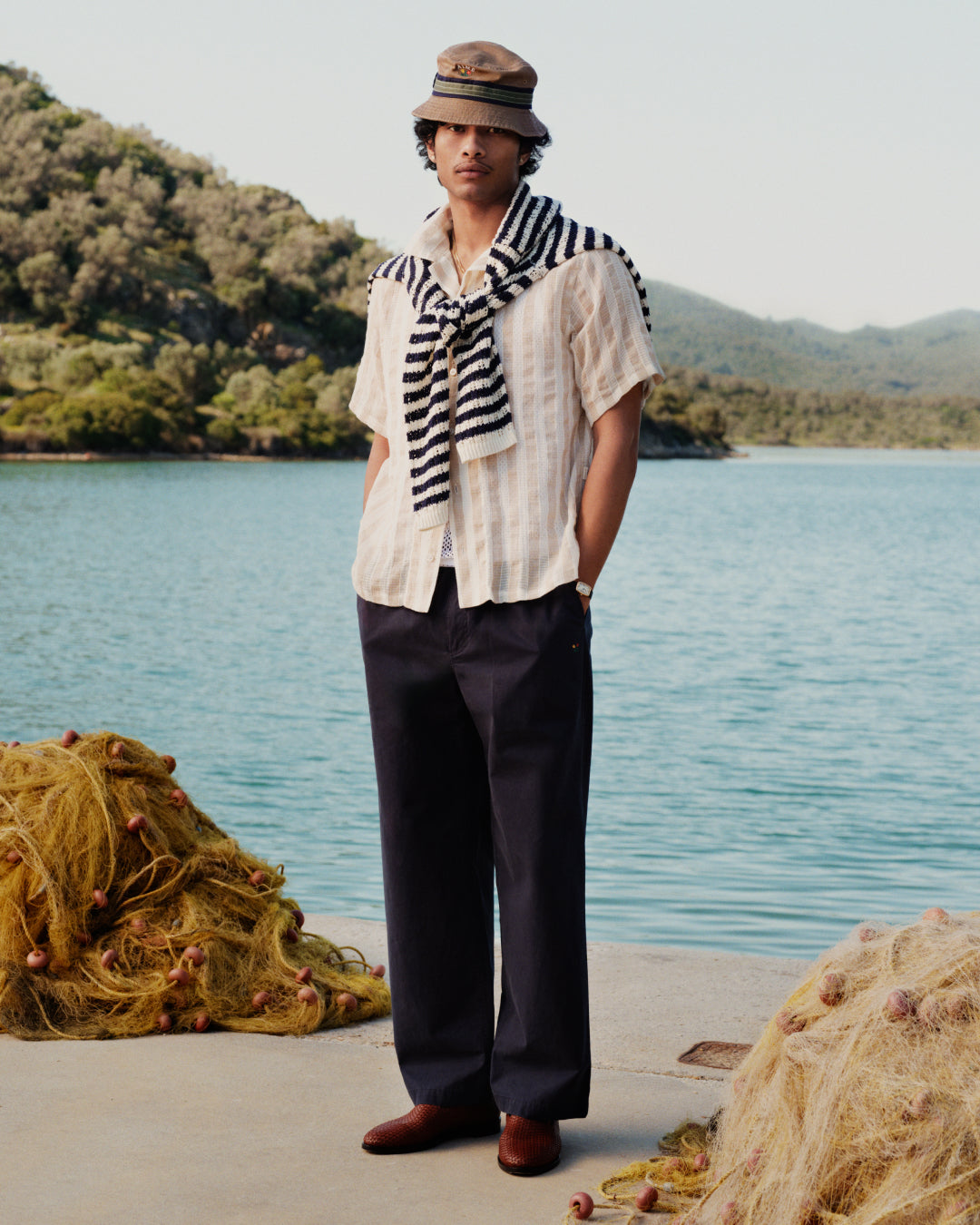 A man by a lake wears a striped sweater, beige hat, white shirt, and Washed Cotton Windbreaker Pant.