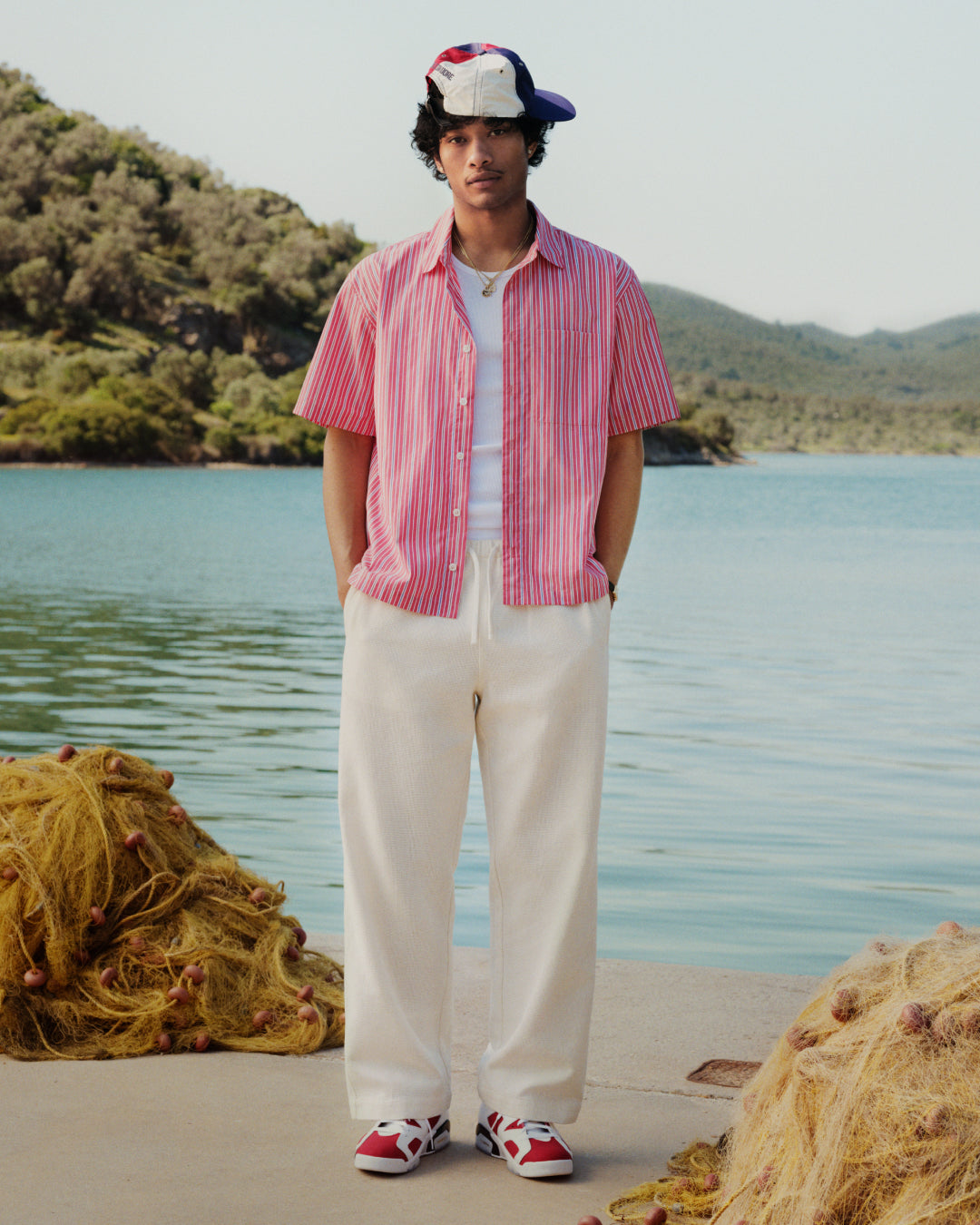 A man in a Yarn Dyed Striped Shirt, white pants, and a multicolored cap stands by water with hills behind him.