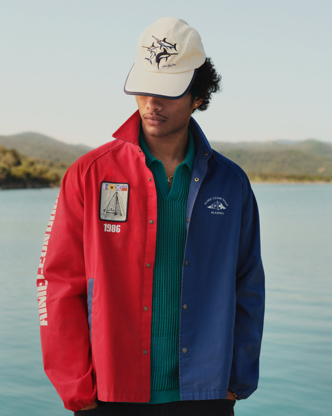 A man in a Marina Jacket and cream hat stands by water with trees and hills in the background.