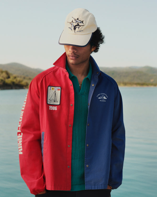 A man in a green Mesh Knit Polo, red and blue jacket, and cream hat stands by water with hills behind him.