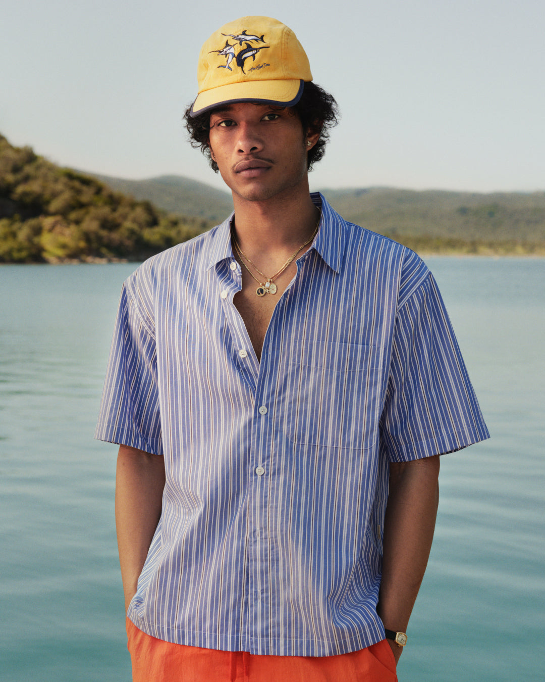 A man in a yellow cap and orange shorts wears a Yarn Dyed Striped Shirt by a lake with hills in the background.