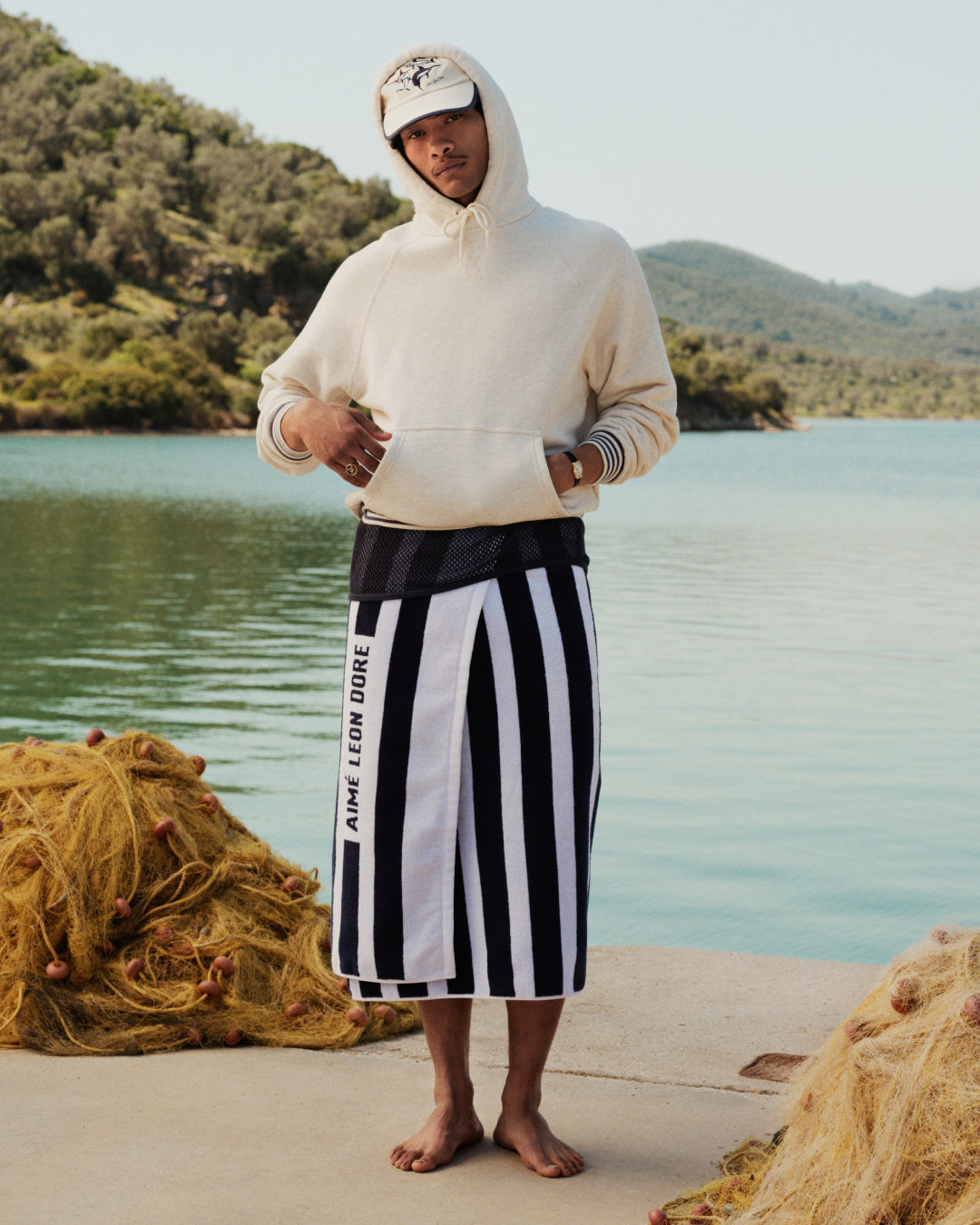 A man in a hoodie, cap, and Striped Beach Towel stands barefoot by a lake with fishing nets and trees behind him.