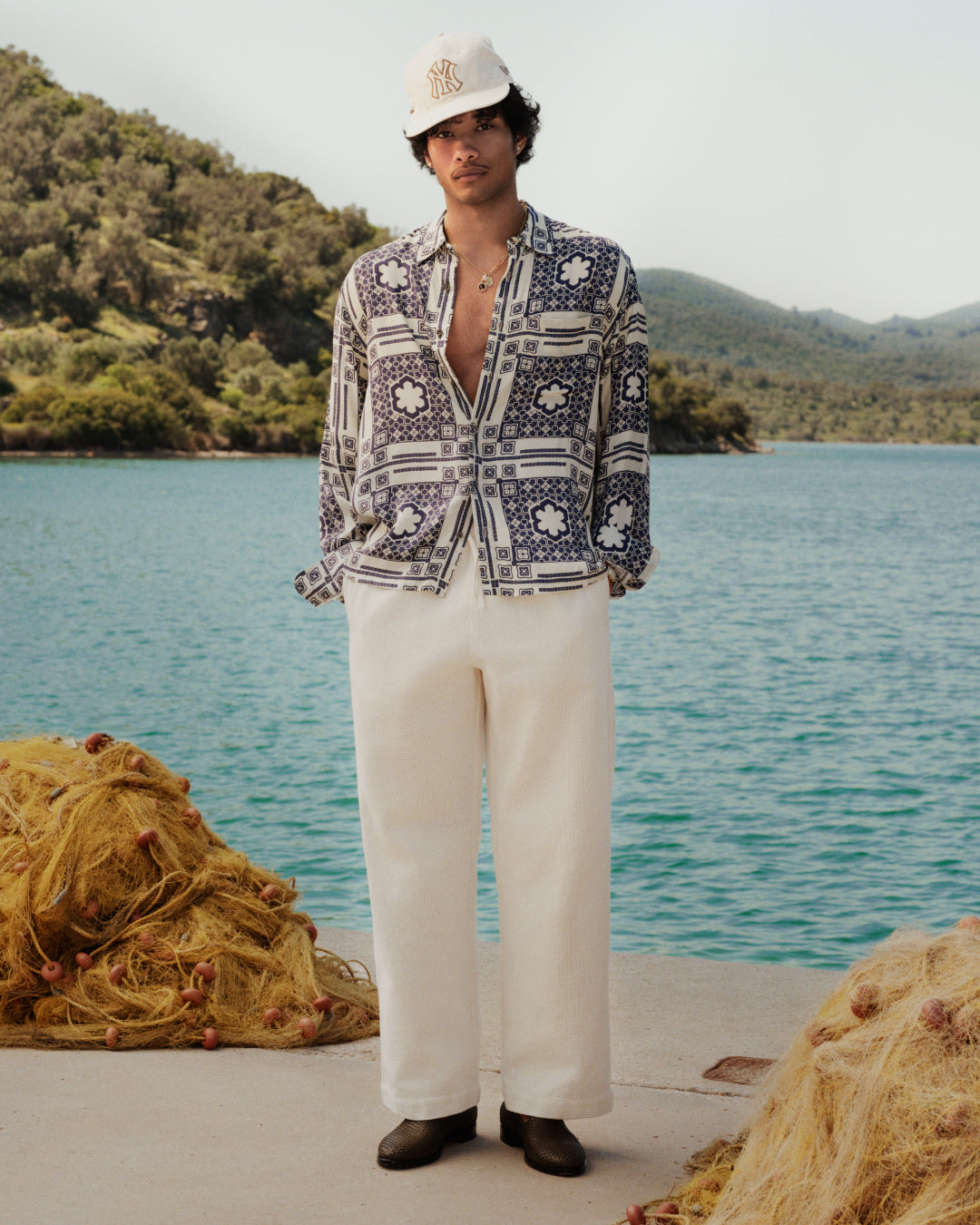 Man in a Mosaic Button Down Shirt, white pants, and cap stands by a lake with nets and hills in the background.