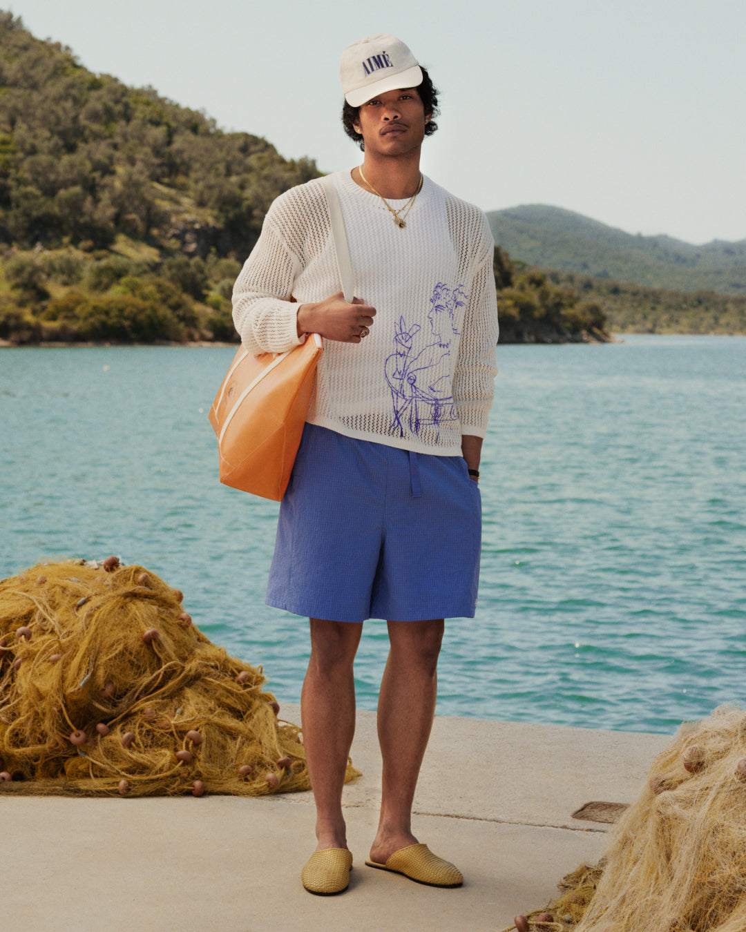 Man in Ripstop Nylon Wave Short, white sweater, and cap stands by water with mountains, nets, and an orange bag.