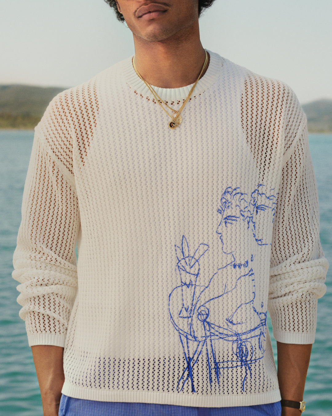 Someone in a Fassianos "Rendez - Vous" Crewneck Sweater with gold necklaces stands by the water.