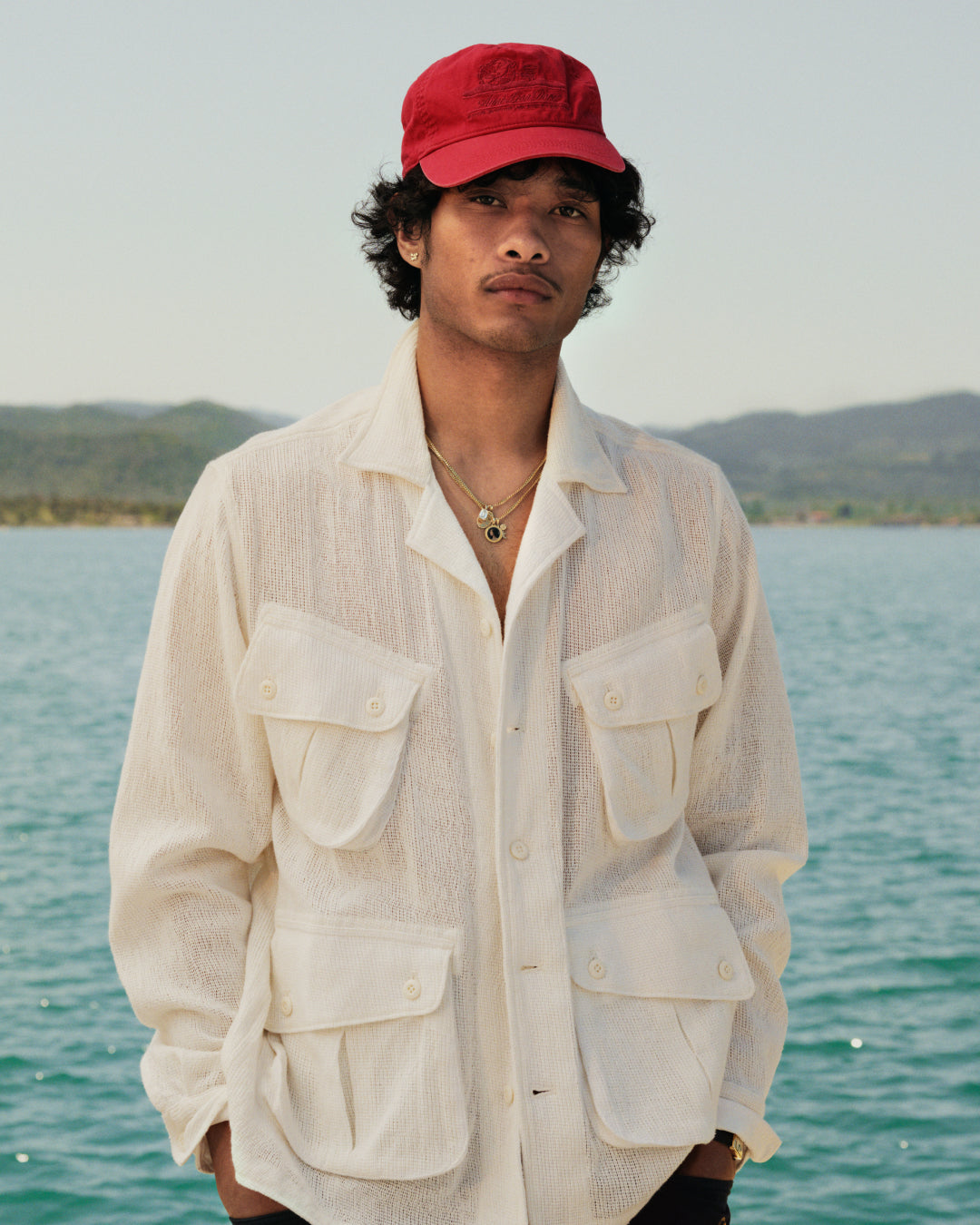 A man in a red cap and Mesh Jungle Shirt stands by blue water with hills in the background.