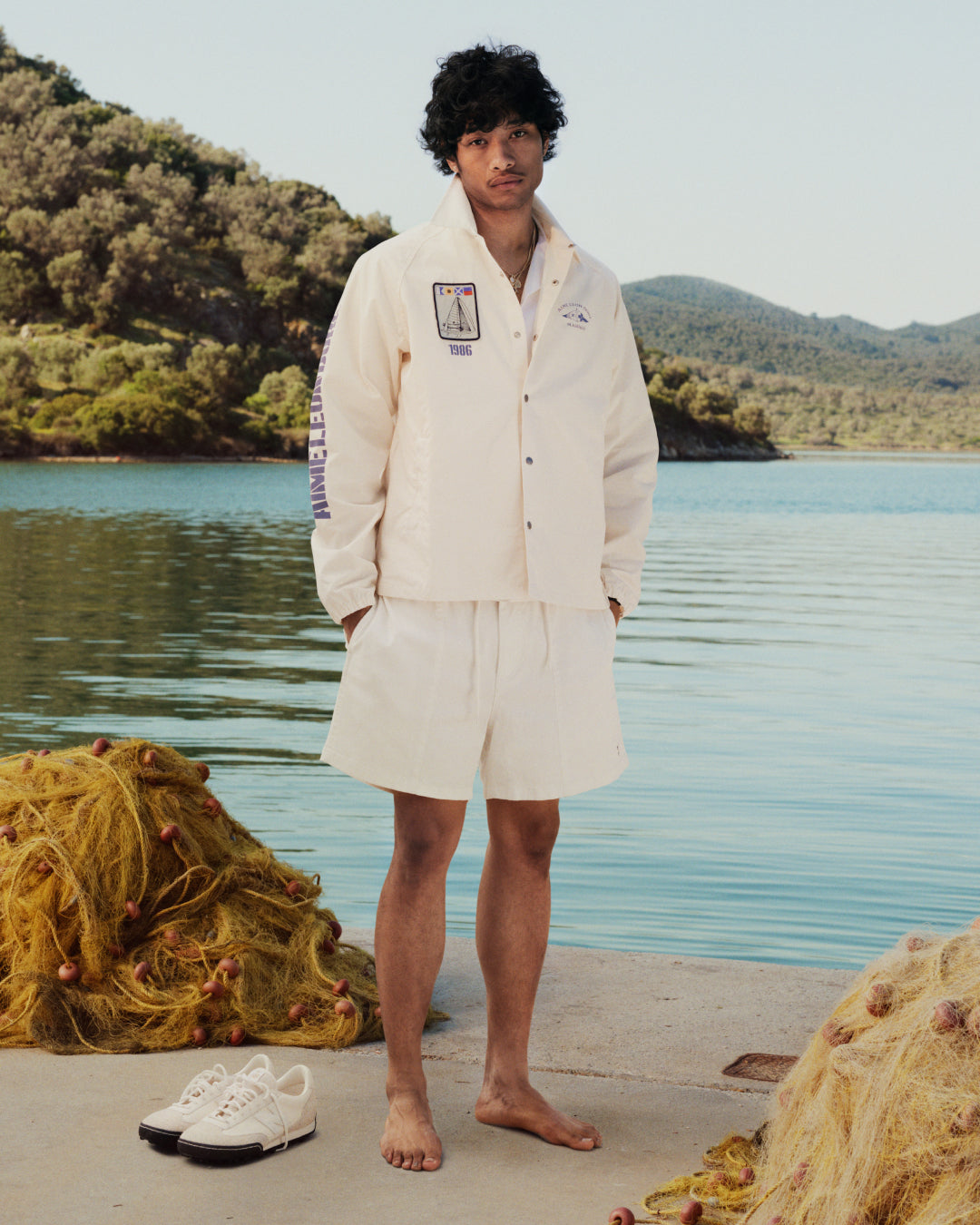 A man in a Marina Jacket and shorts stands barefoot on a dock by a lake, with shoes and fishing nets nearby.