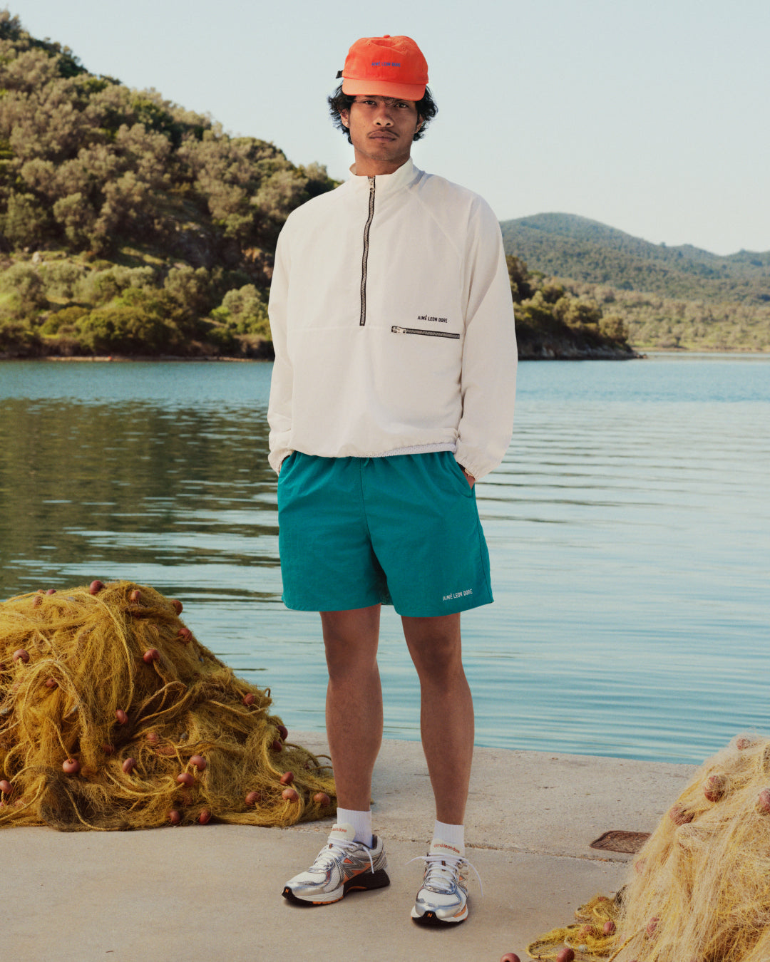 A person in ALD / New Balance 860v2 shoes stands by a lake with mountains and fishing nets in the background.