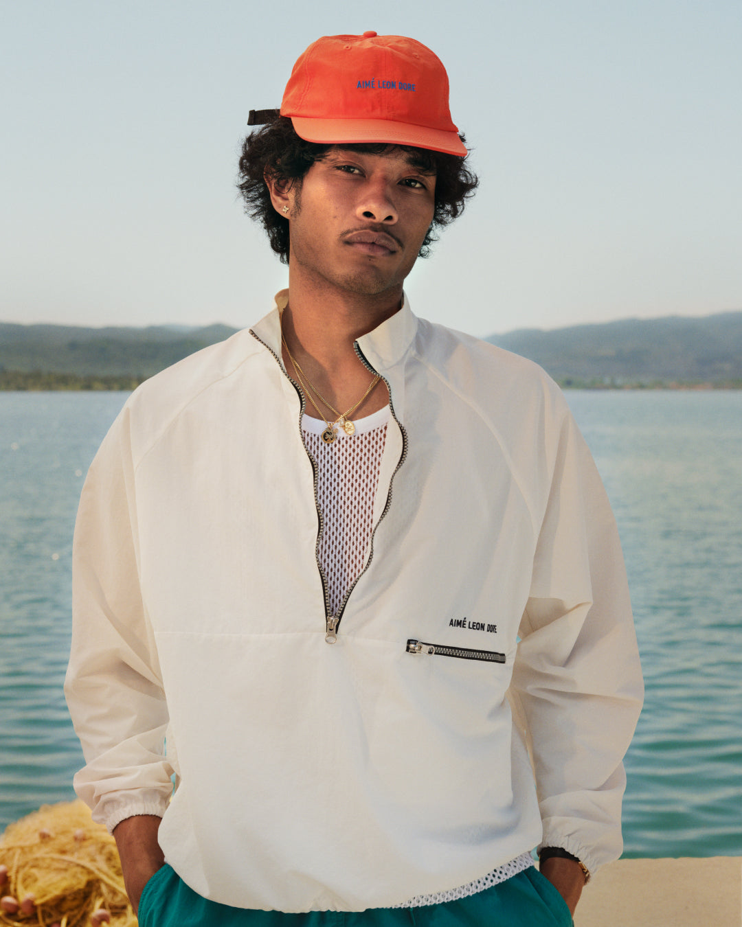A person in a Nylon Packable Windbreaker and orange cap stands by the water with mountains in the background.