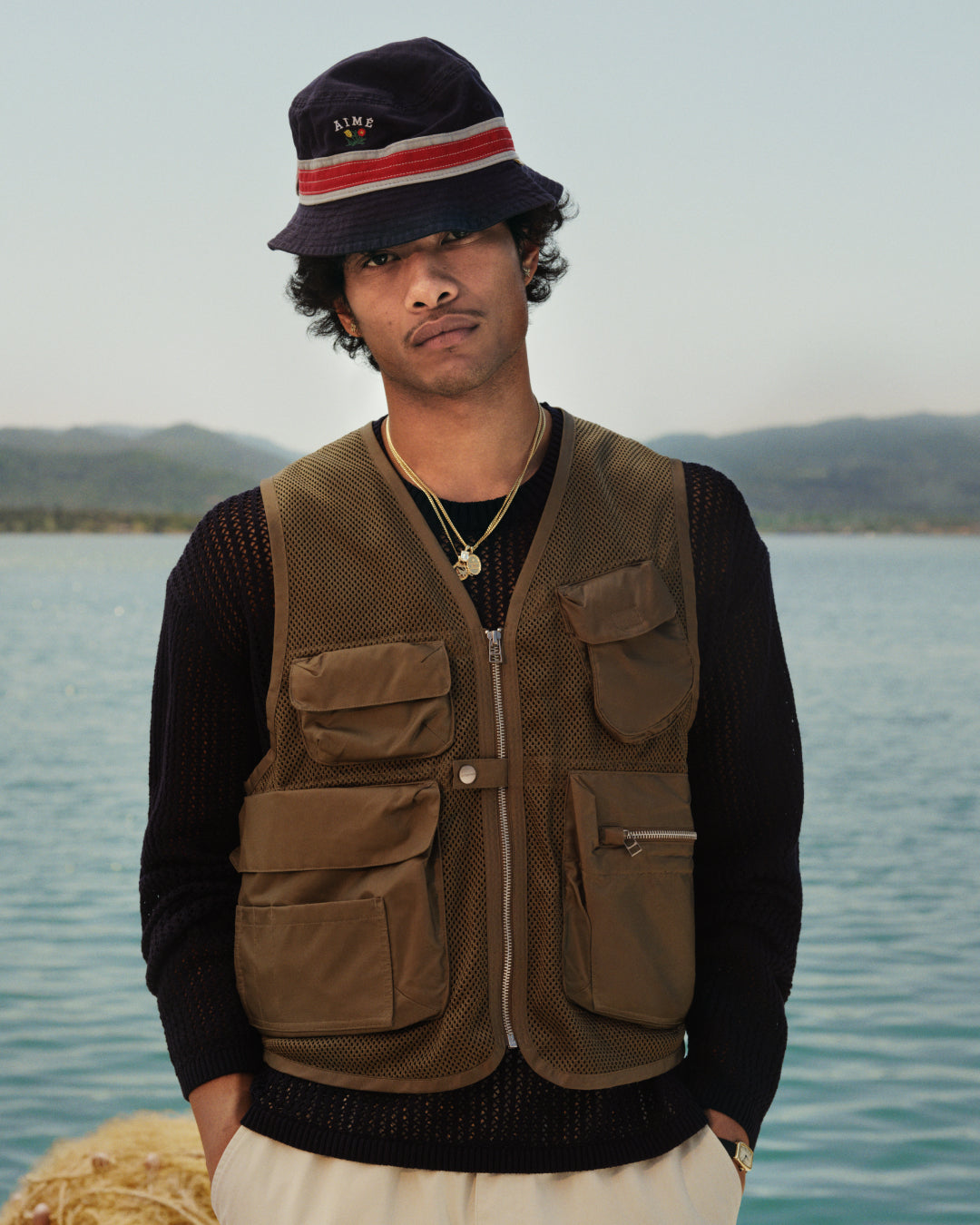A man in a Nylon Utility Vest and bucket hat stands by a lake with mountains in the background.