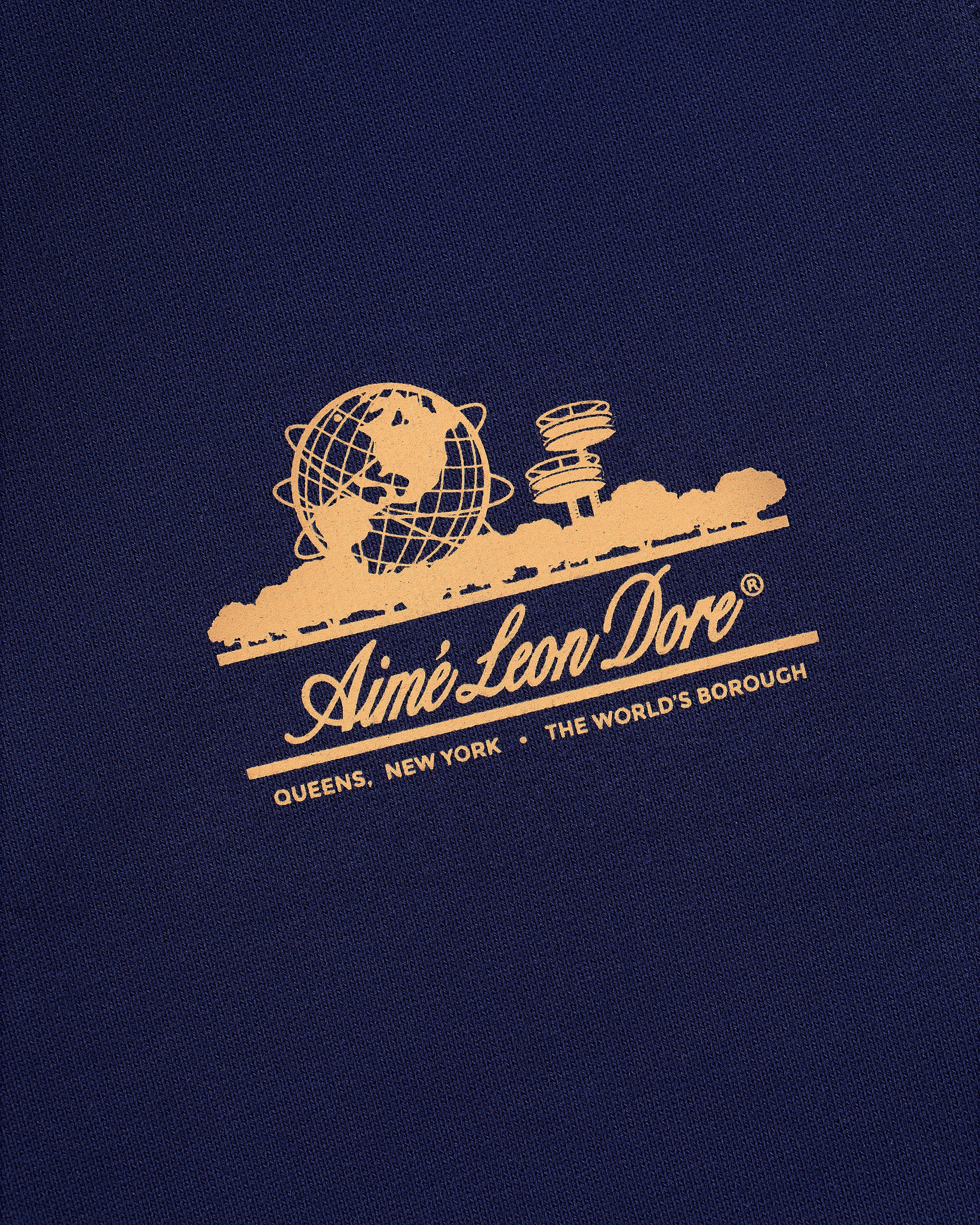 Gold Aimé Leon Dore logo featuring globe and skyline on dark blue Unisphere Sweatpants; reads Queens, New York.
