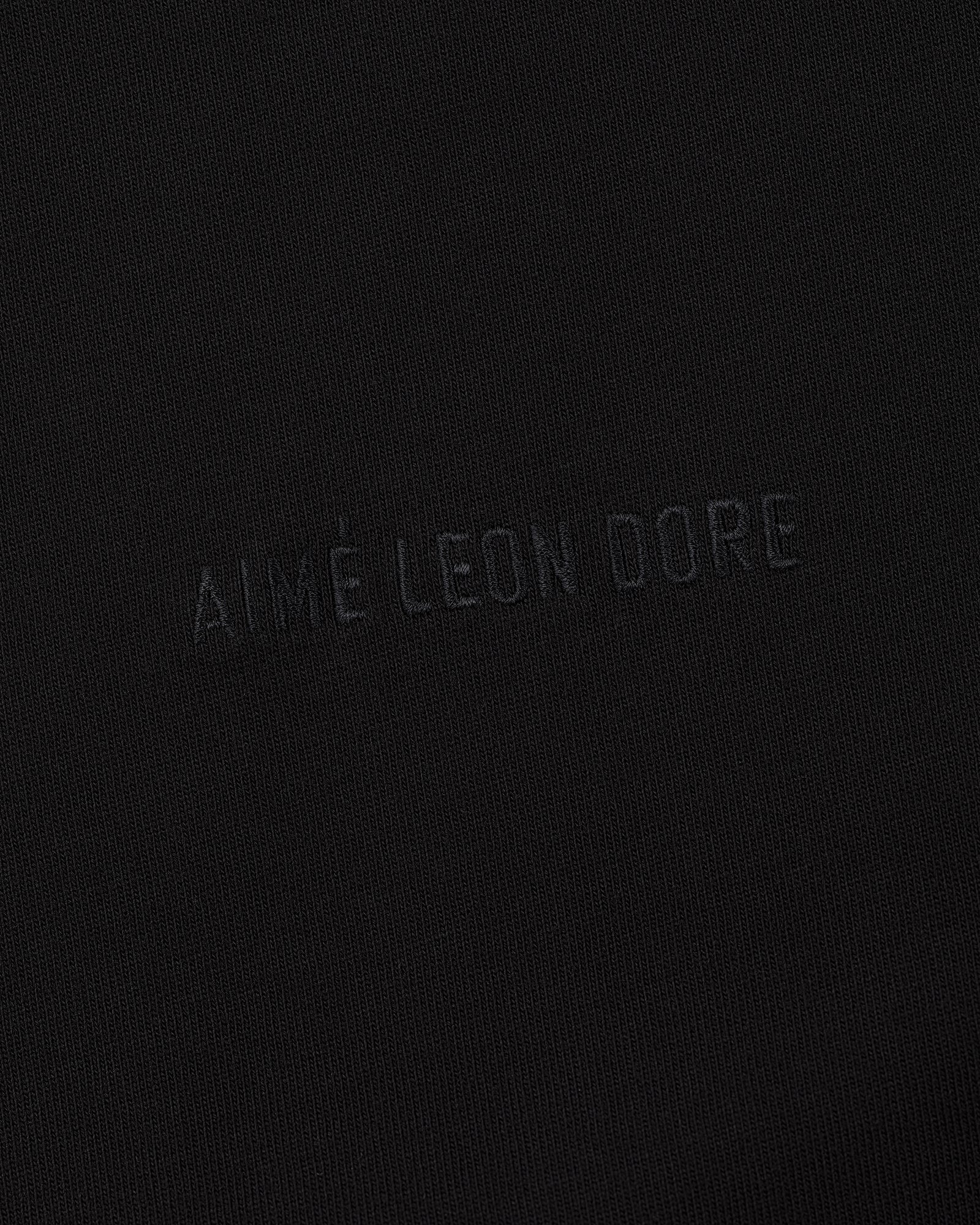 Close-up of Tonal Logo Tee's black fabric with AIMÉ LEON DORE subtly embossed in matching black lettering.