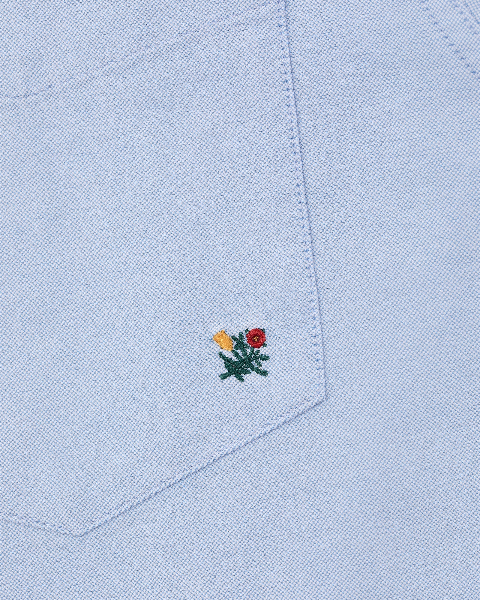 Light blue Crest Oxford Shirt with a fabric pocket featuring a small red and yellow flower embroidered near the bottom.