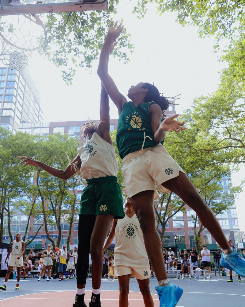 Two women playing basketball on an outdoor court with trees and buildings in the background.