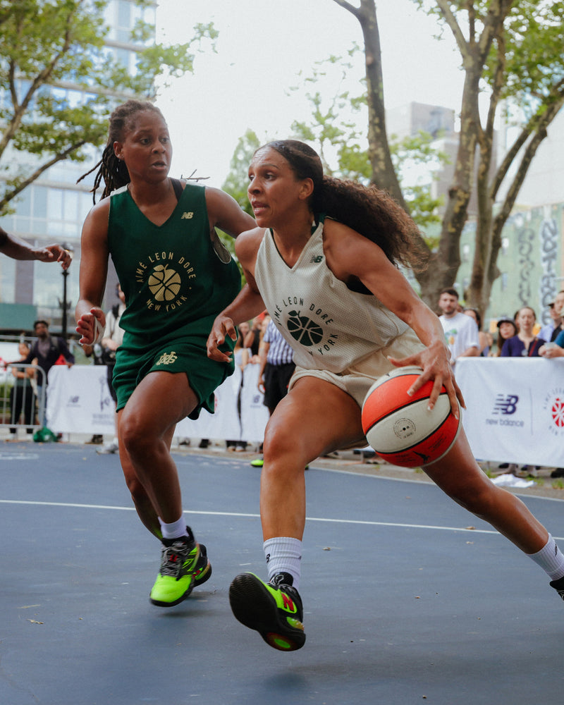 Two women playing basketball on an outdoor court with trees and spectators in the background.