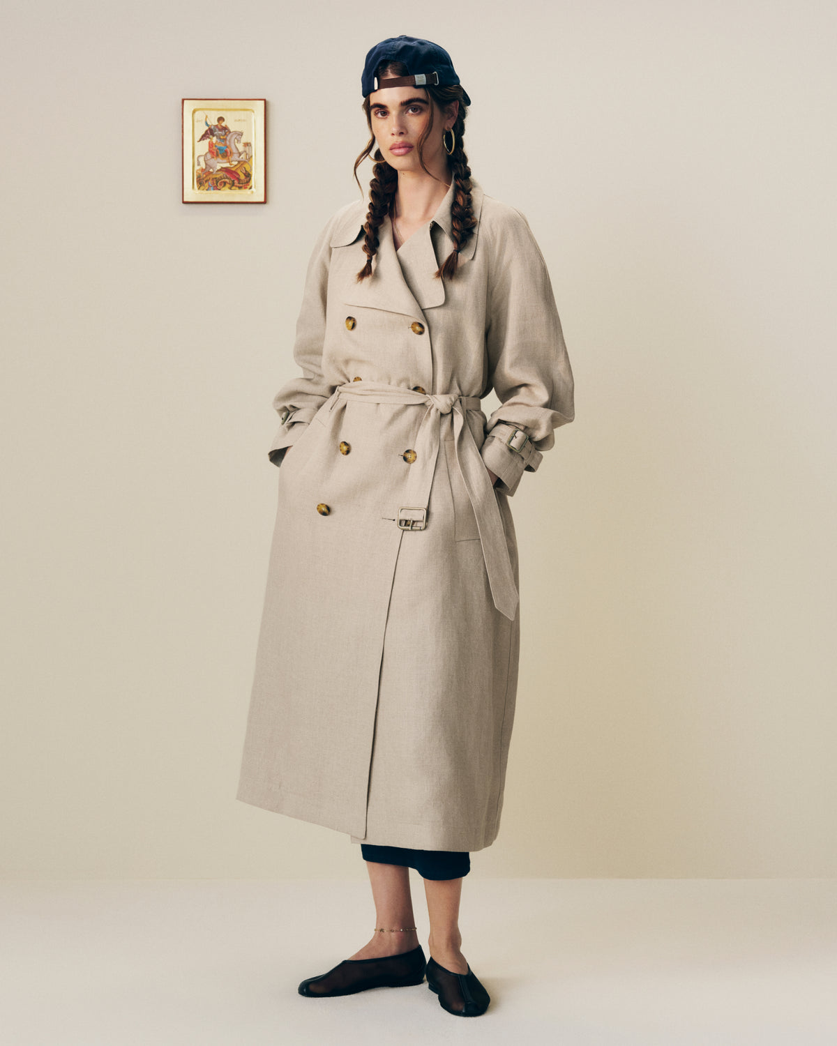 A woman models a Linen Trench Coat, hands in pockets, standing by a plain wall with a small framed picture.