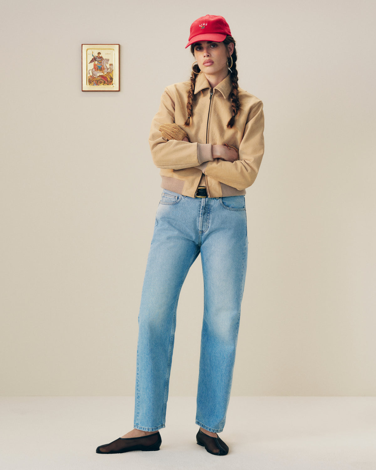 Woman in a Croc-Embossed Nubuck Bomber Jacket, blue jeans, red cap, and black flats stands indoors by a small picture.