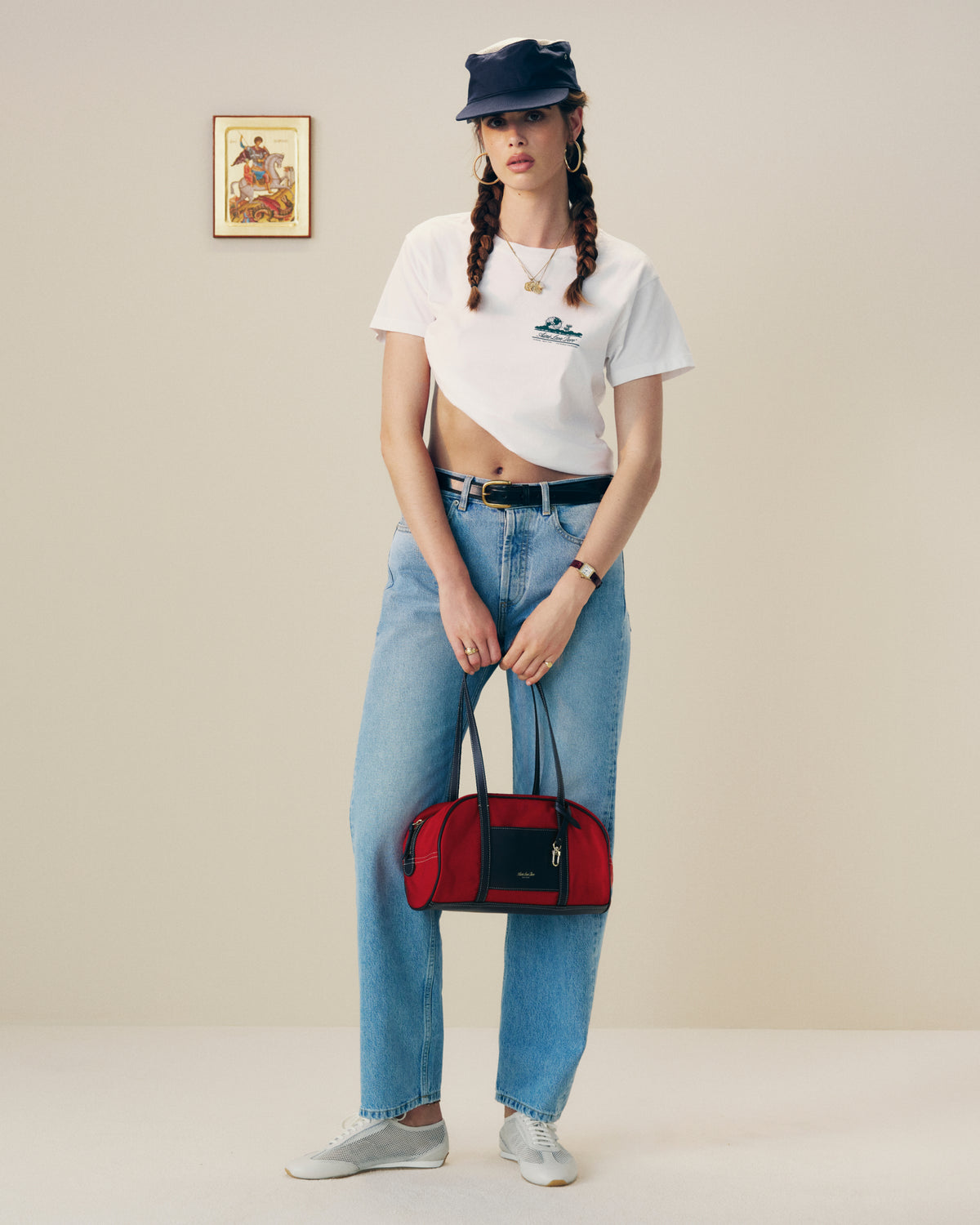 Woman in a white crop tee, jeans, and cap holds the Mini Duffle Shoulder Bag in red, standing by wall art.