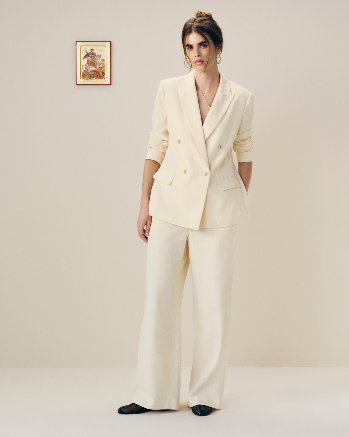 A woman models Drawstring Cotton Blend Suit Trousers in cream against a plain wall with framed art behind her.